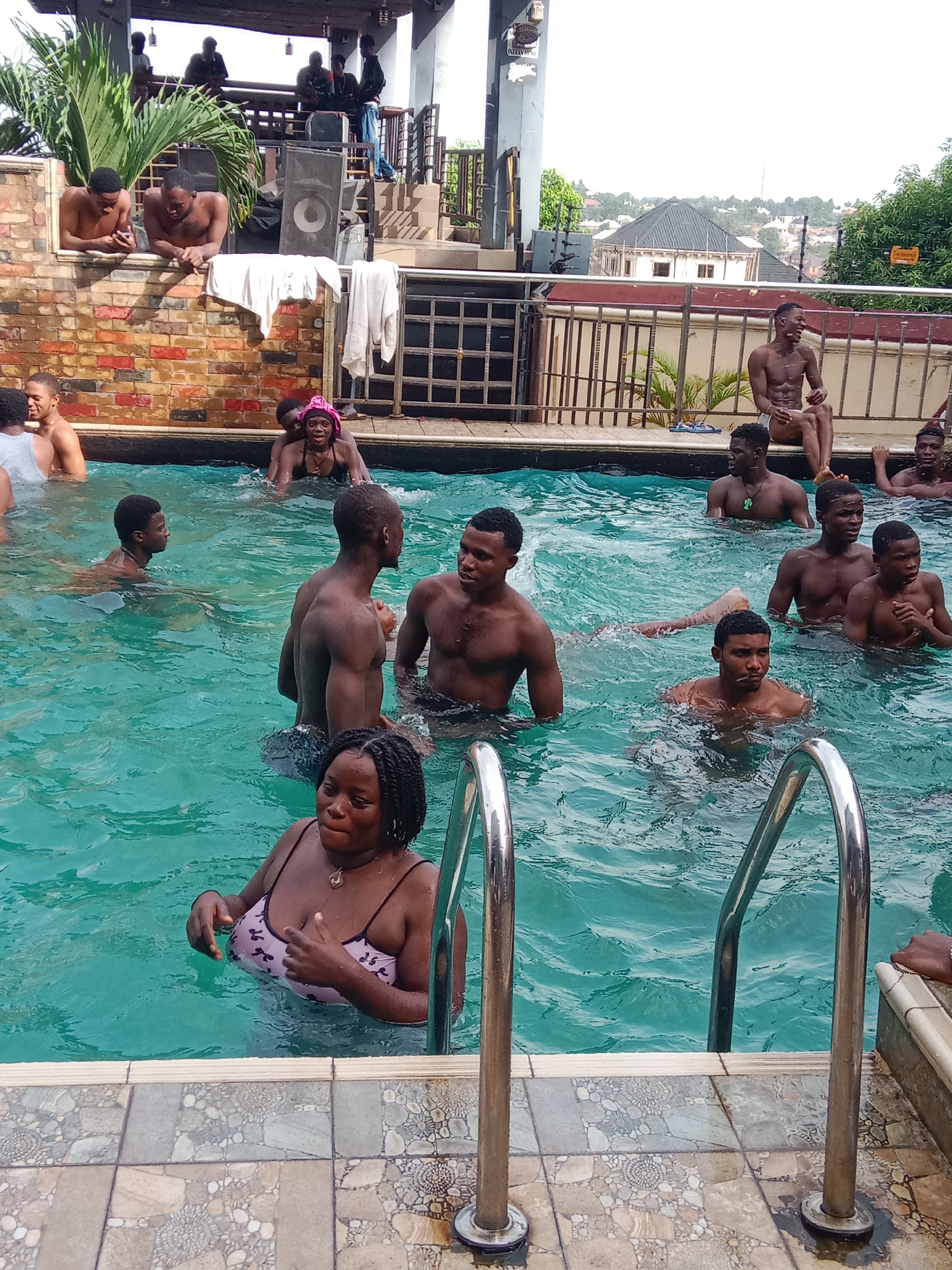 A partially outdoor swimming pool scene in Onitsha, Nigeria is captured. Individuals of African descent are seen both in and around the pool. Some people are in the water, swimming, standing, or conversing. Others are sitting or standing poolside. One woman is near a metal pool ladder. A brick wall and a second level structure are in the background, complete with people and audio equipment. The surrounding area reveals foliage and distant buildings, suggesting an urban or suburban setting.
