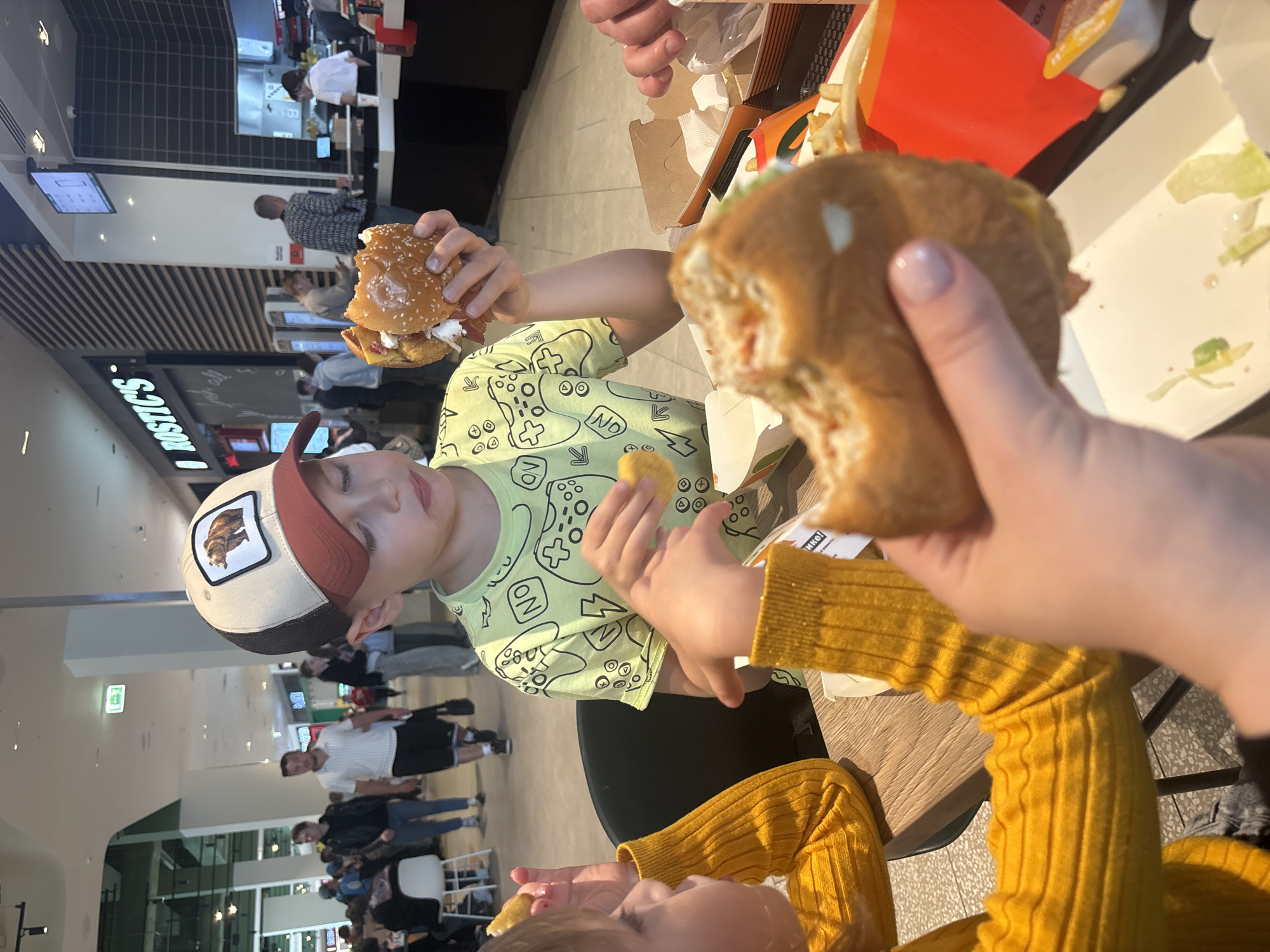The scene depicts individuals eating fast food within a brightly lit food court, identified by the prominent "ROSTIC'S" sign, located in Perm, Russia. In the immediate foreground, a child, likely female, wearing a yellow ribbed long-sleeve sweater, is visible from the left, holding a golden-brown chicken nugget in her left hand. To her right, a young boy, approximately 6-10 years old, wears a light green short-sleeve t-shirt patterned with black outlines of video game controllers and "ON" text. He wears a beige and brown baseball cap featuring a square patch with a brown bear illustration. He holds a partially consumed hamburger with a sesame seed bun in both hands, positioned near his mouth.
Further in the foreground, at the bottom right, an adult hand with light pink nail polish is visible, holding another partially eaten hamburger. On the wooden table, multiple fast-food items are present, including French fries in an orange container, additional chicken nuggets, and open white cardboard boxes containing burger remnants, including lettuce and sauces.
The background reveals a bustling shopping mall environment. The Rostic's establishment features black horizontally slatted walls, vertical black tiled sections, and digital menu screens above the service counter. Several self-service kiosks are visible near the entrance. Behind the counter, a staff member in a white uniform shirt and dark trousers attends to the service area. Numerous individuals are walking throughout the wider food court area; notable figures include a man in a white polo shirt and dark shorts carrying a black bag, and another man in a black leather jacket and blue jeans. The floor is light-colored, and the ceiling is high with modern lighting fixtures.