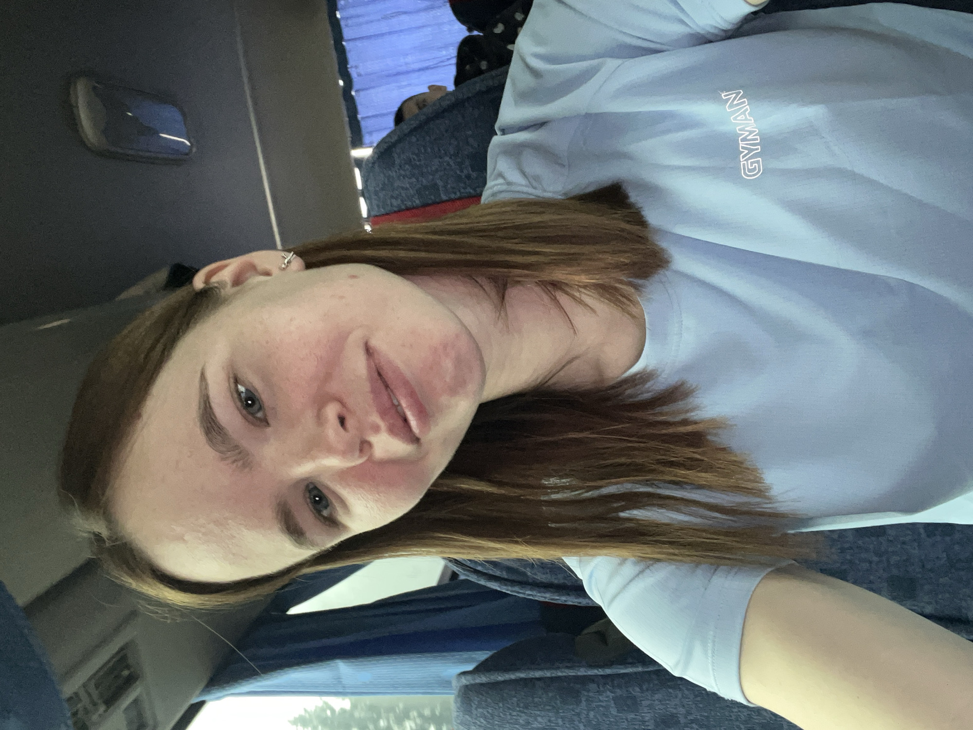 A woman is taking a selfie on what appears to be a bus. She has brown hair, fair skin, and is wearing a light blue t-shirt with the word "GYMAN" printed vertically on the chest. The bus has blue seats and a window. The location is unknown, I could not find Большая Уса, Russia to have a cityscape from which to compare to.