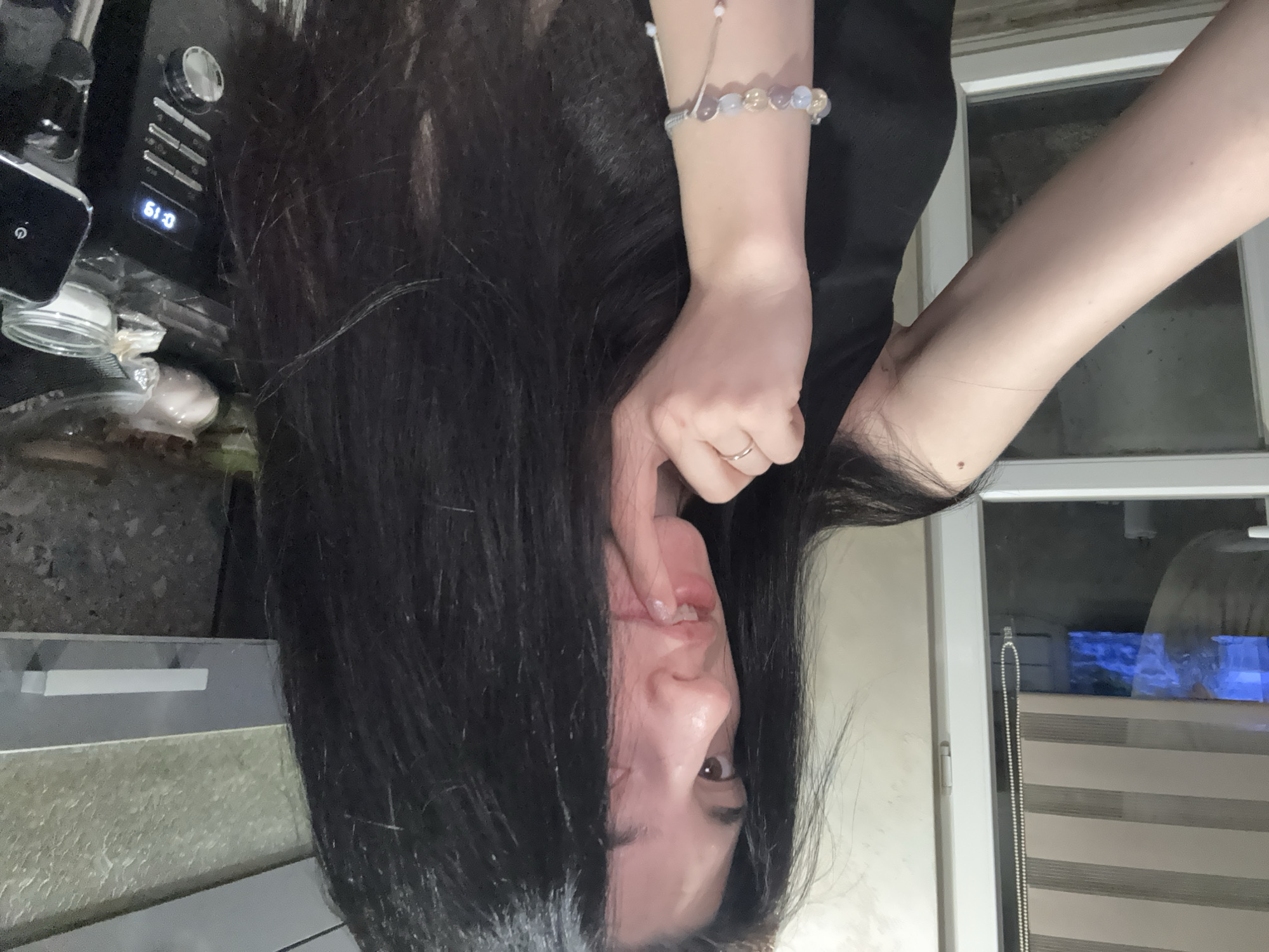 A woman with long black hair is upside down, holding her lip with her index finger and thumb. She is wearing a black top and a beaded bracelet on her wrist. Behind her are kitchen appliances and a window with partially closed blinds. The location is not identifiable as Sofia, Bulgaria based on the image content.