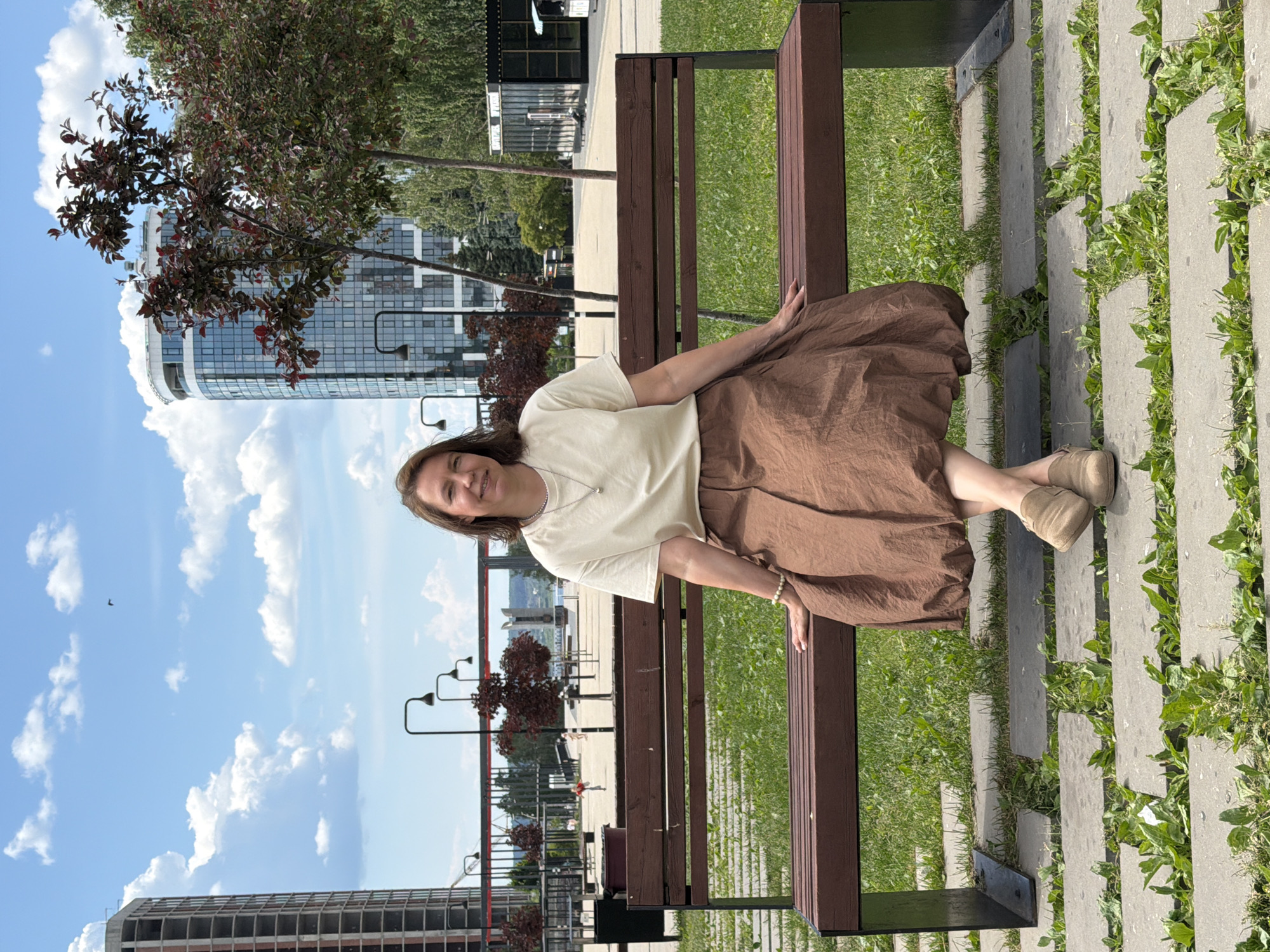In Izhevsk, Russia, a woman sits on a park bench with a brown wooden seat and dark metal legs. She is wearing a light-colored t-shirt, a voluminous brown skirt, and light-brown shoes. The bench is situated on a paved pathway with grass growing between the stones. In the background, two modern high-rise buildings are visible, one under construction and another with a curved glass facade. Trees with dark red leaves are also present, along with streetlights and other urban elements, under a bright blue sky with white clouds.