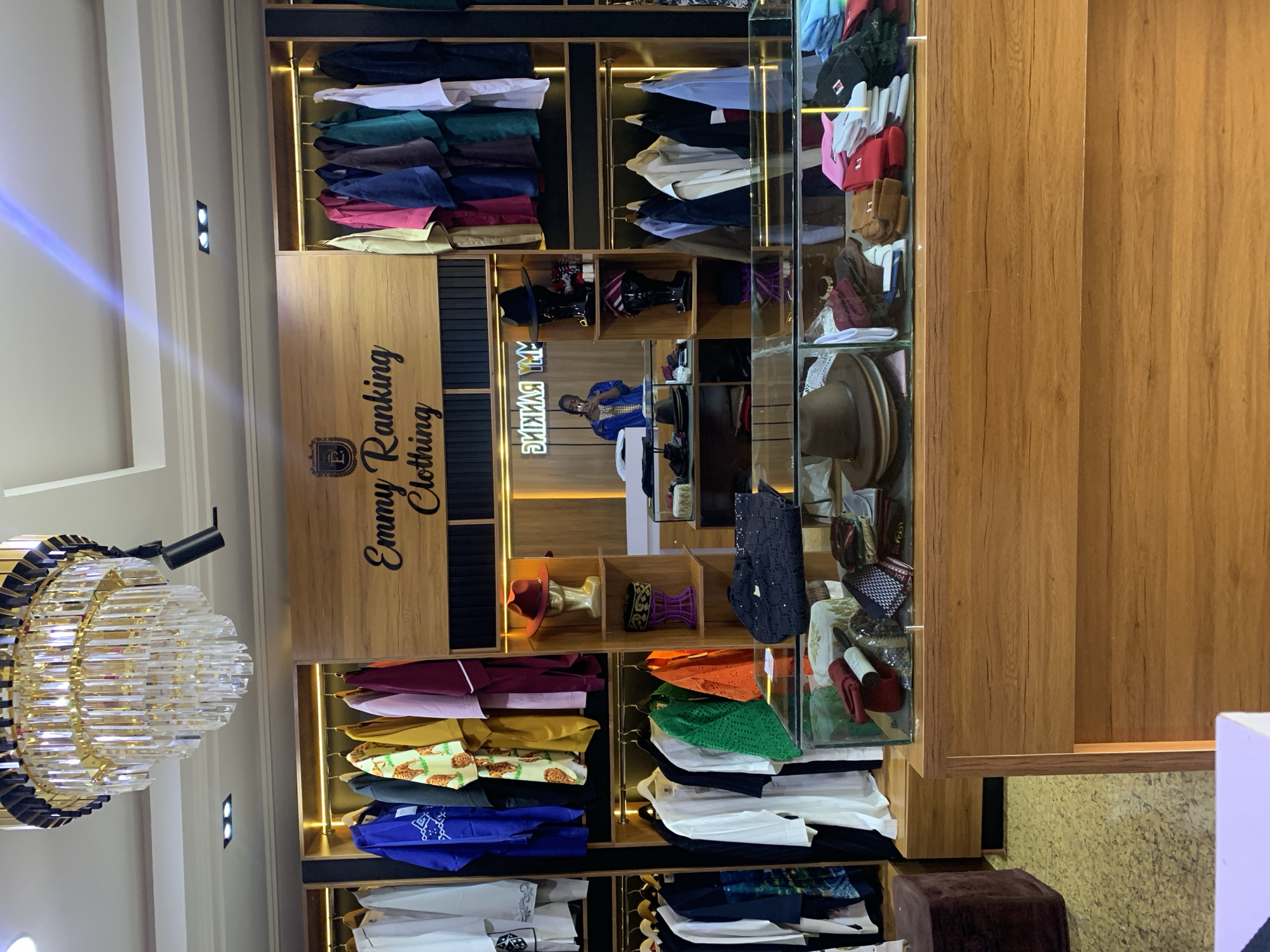 This is a photograph of the interior of "Emmy Ranking Clothing," a retail store located in Gura Top, Nigeria. The store features a well-lit and organized display of clothing, hats, and accessories.
In the foreground, a polished wooden counter with a glass display case is visible. The display case contains a variety of hats, some folded neatly, others stacked. Visible items include what appear to be fedoras and possibly some skullcaps. Also in the display are folded fabrics and what seem to be small accessories like ties or scarves.
Behind the counter, the store's display walls are lined with shelving and hanging racks. On the left side, several white and black garments with embroidered details are hung. Further into the store, more clothing is displayed on hanging racks, with items ranging in color from blue and yellow to maroon and orange. A mannequin head wearing a red hat is displayed on a shelf.
The back wall features the store's name, "Emmy Ranking Clothing," in a stylized font against a wooden panel. A woman, presumably an employee, is standing behind a counter in the background, taking a photograph with her phone. She is wearing a blue jacket over a lighter garment.
The store's ceiling is adorned with a large, multi-tiered crystal chandelier. Recessed lighting fixtures are also present on the ceiling. The floor appears to be covered with a textured, speckled material. The overall impression is of a modern and well-maintained boutique.