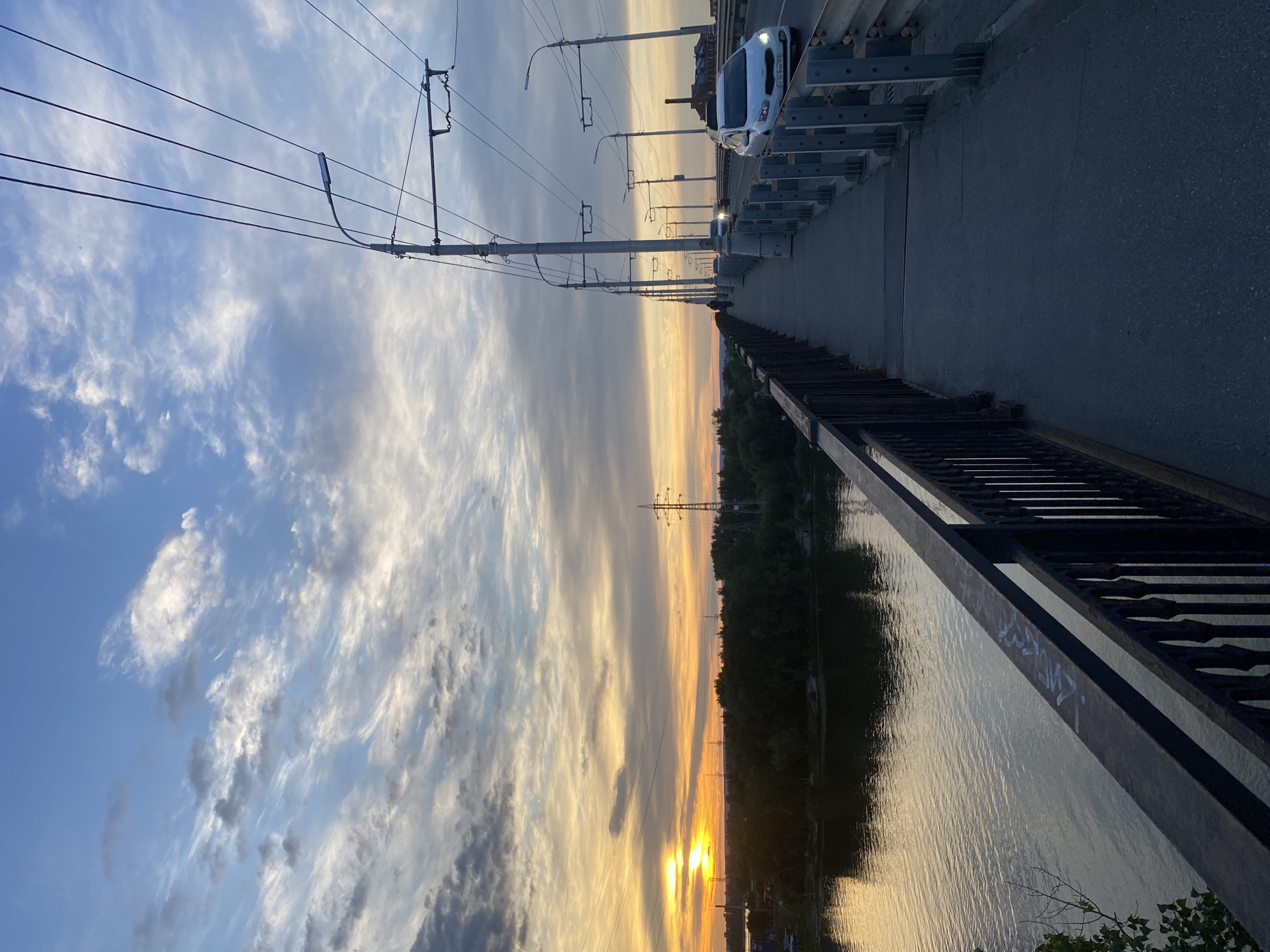 The image depicts a scenic view from a bridge, likely during sunset, given the warm hues of the sky. The scene features a roadway with a white car and overhead electric wires, suggesting a transportation route. To the right, a body of water reflects the sky and the surrounding trees, which form a dense line along the bank. A metal railing runs along the edge of the bridge, and some graffiti is visible on its surface. In the distance, power lines and towers are seen against the colorful sky, hinting at the presence of industrial infrastructure. The overall mood is tranquil, enhanced by the natural beauty of the sunset and the calm water. The location context provided is Novokuybyshevsk, Russia.