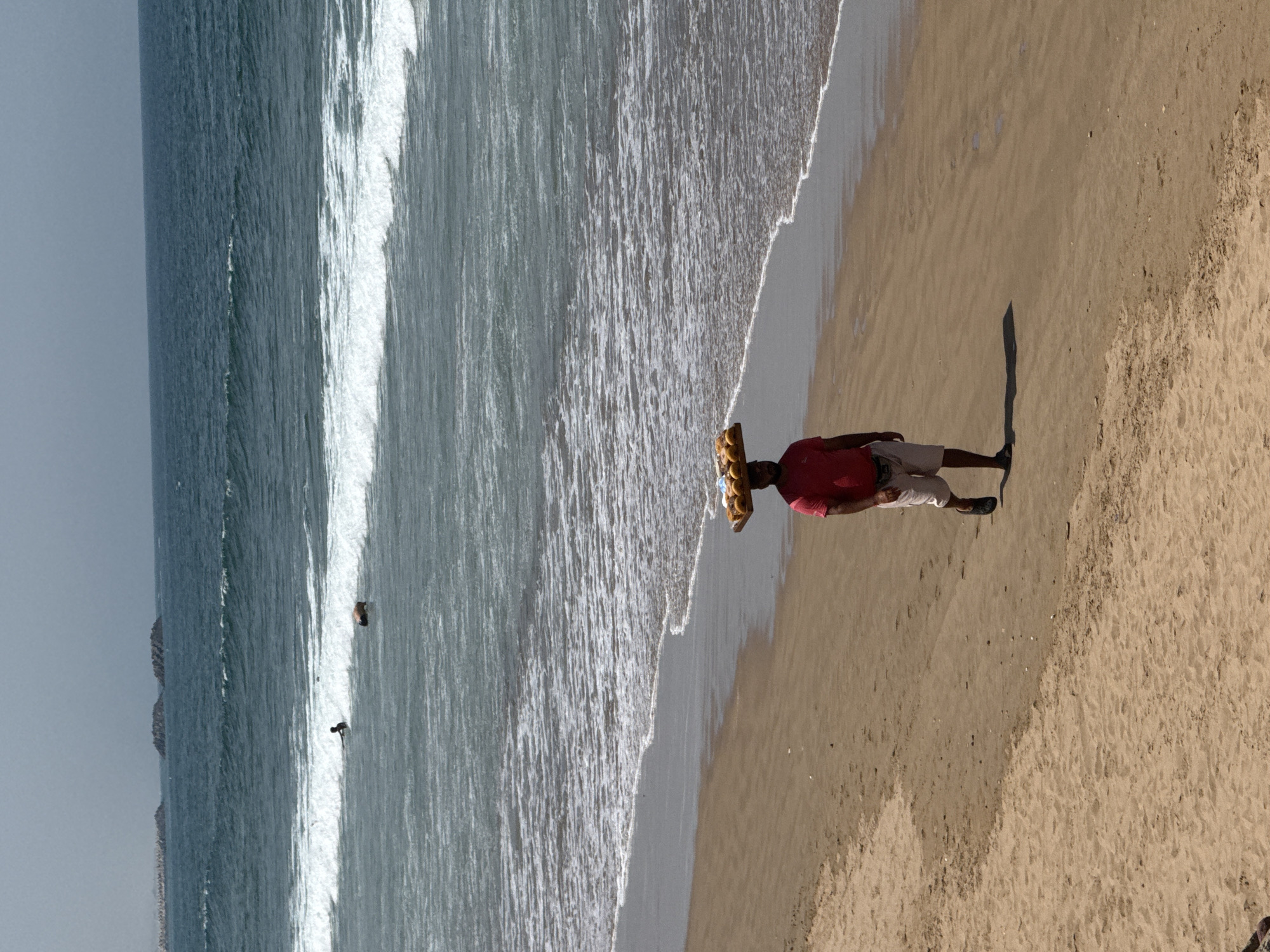 A man wearing a red shirt and light-colored pants walks along a beach in Agadir, Morocco. He carries goods stacked on his head. The beach is sandy with waves approaching the shoreline. In the distance, two people are in the ocean. The sky is overcast.