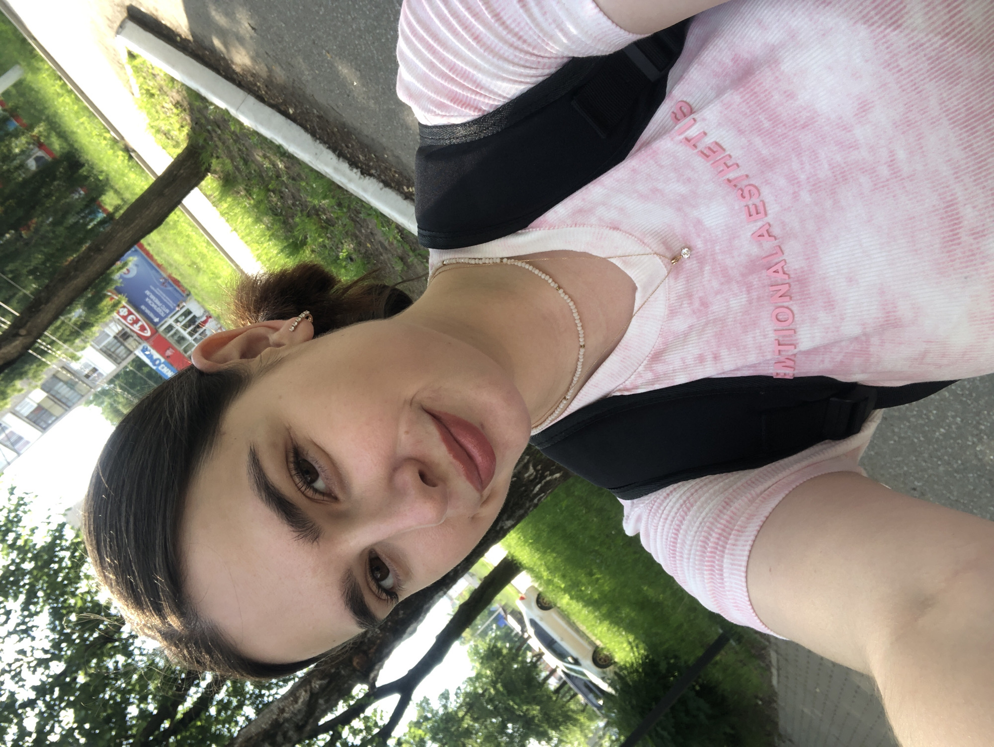 The image is a close-up selfie of a young woman with dark hair, brown eyes, and light skin. She is wearing a pink and white tie-dye t-shirt with the text "EMOTIONAL AESTHETIC" in pink letters across the chest, a delicate necklace with a small pendant, and a black backpack. Her hair is pulled back, and she is wearing small hoop earrings. She has makeup on, including lipstick. The background suggests an outdoor urban environment, with trees, grass, a sidewalk, and a road. In the background, there are buildings and a parked car. The lighting indicates it is daytime. Given the provided context of Chaikovsky, Russia, the scene is likely a street or park area within the city.