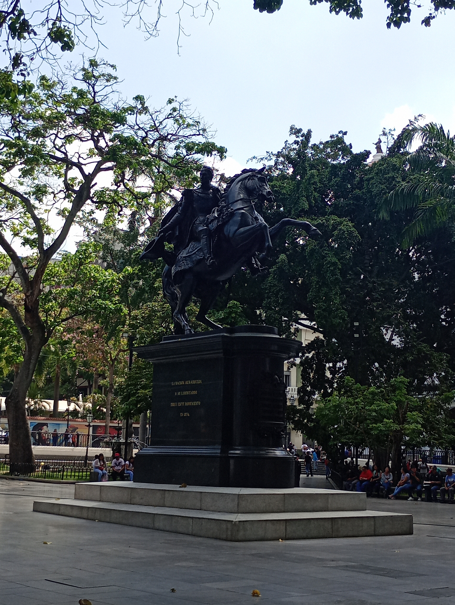 The image shows a statue of Simón Bolívar on horseback in Plaza Bolívar, Caracas, Venezuela. The statue is dark in color and stands on a large rectangular pedestal. The pedestal has an inscription, which reads "LA NACION AGRADECIDA / A SU LIBERTADOR / ERIOSE ESTE MONUMENTO / EN 1874." Trees surround the statue, and people are visible sitting on benches in the background. The sky is partly cloudy. The ground is paved with stone or concrete.