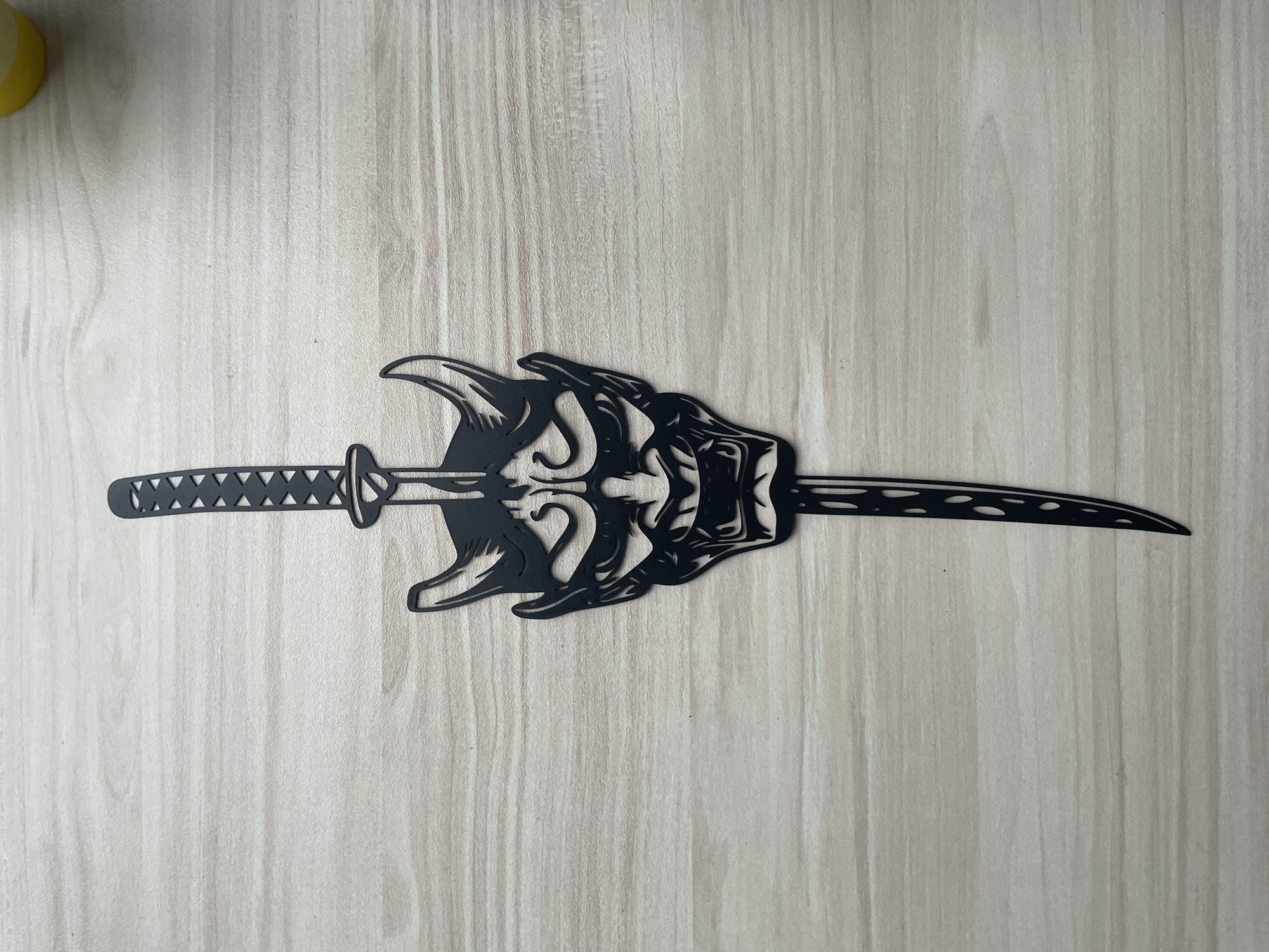 A katana, held in a stylized hand, lies on a light-colored wooden surface. The katana is black and ornate. The hand is similarly stylized and appears to be grasping the blade. Part of a yellow object is in the upper-left corner. The location is unknown.