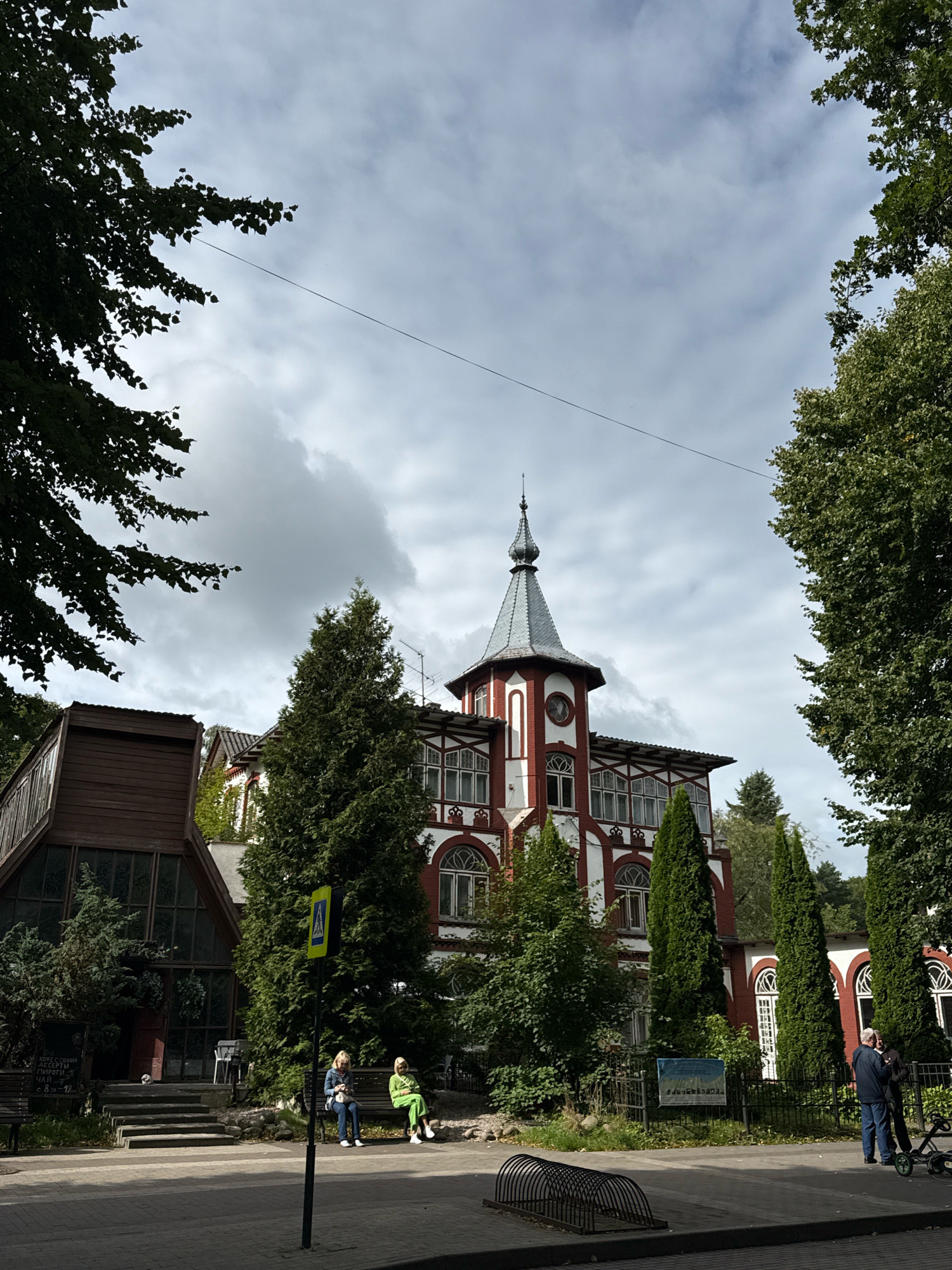 In Svetlogorsk, Russia, a red and white multi-story building with a prominent tower and arched windows stands amidst lush greenery. The architecture suggests a historic or whimsical design. In front of the building, two women are seated on a bench, one in a blue jacket and the other in a bright green outfit. Adjacent to them, a yellow pedestrian crossing sign is visible. To the left, a rustic, wooden structure with large glass windows is partially obscured by trees. Further to the right, two men are standing near a fence, one dressed in a dark jacket and the other in a blue one. A stroller is parked nearby. The scene is under a cloudy sky, with trees framing the view.
