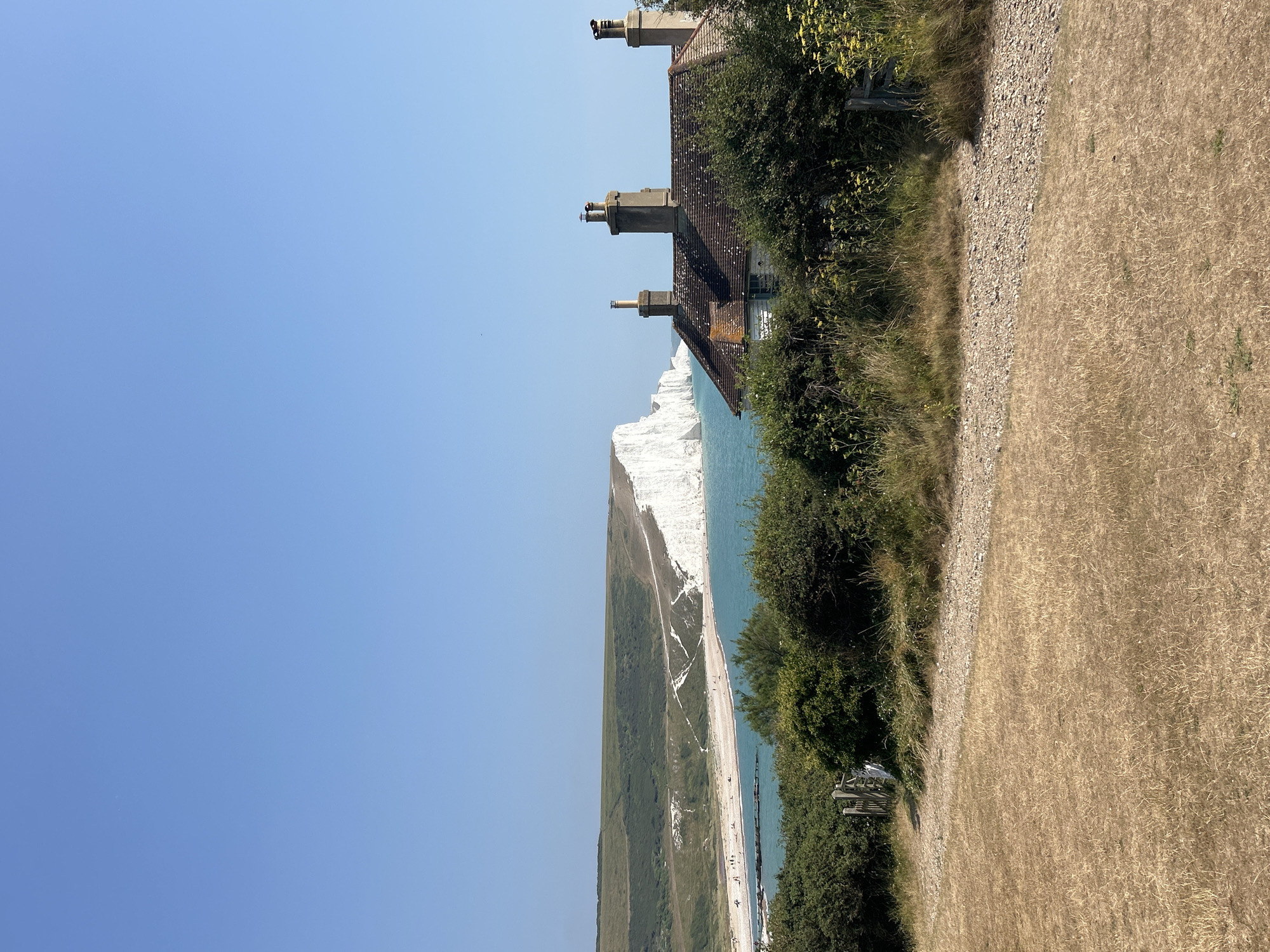 This image shows a view from a hillside towards the Seven Sisters cliffs in Seaford, United Kingdom. In the foreground, dry, tan-colored grass covers the ground, with a gravel path leading through it. A wooden gate is visible on the left. Behind the gate and path, dense green bushes and yellow wildflowers grow. Further back, a small, weathered house with a tiled roof and three chimneys sits nestled amongst trees. Beyond the house, the turquoise sea meets the iconic white chalk cliffs of the Seven Sisters. A beach with a few scattered people is visible at the base of the cliffs. The sky is a clear, bright blue.