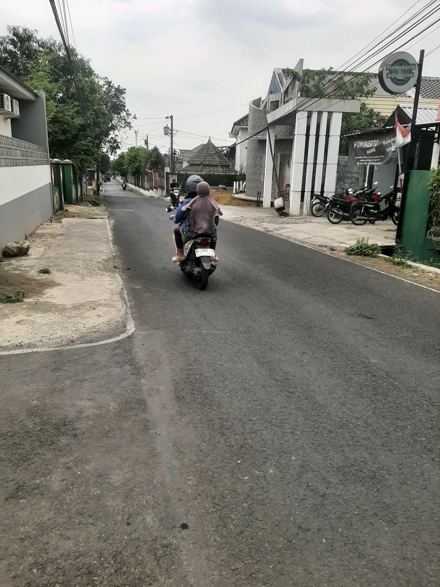 The image captures a view of a street in Sumampir, Indonesia, on an overcast day. In the foreground, the asphalt road is visible, with a concrete sidewalk to the left. A person wearing a blue shirt and a headscarf is riding on the back of a motorcycle, which has a license plate reading "AD 2868 TO". The motorcycle is moving away from the camera. To the right of the road, there is a building with a modern architectural design featuring white pillars and a curved facade, and a circular sign with text that appears to say "Purwokerto & Adventure". Several motorcycles are parked in front of this building, along with a black banner with white text and a small Indonesian flag. Further down the street, more buildings and trees line both sides. The sky is uniformly grey, suggesting cloudy weather.
