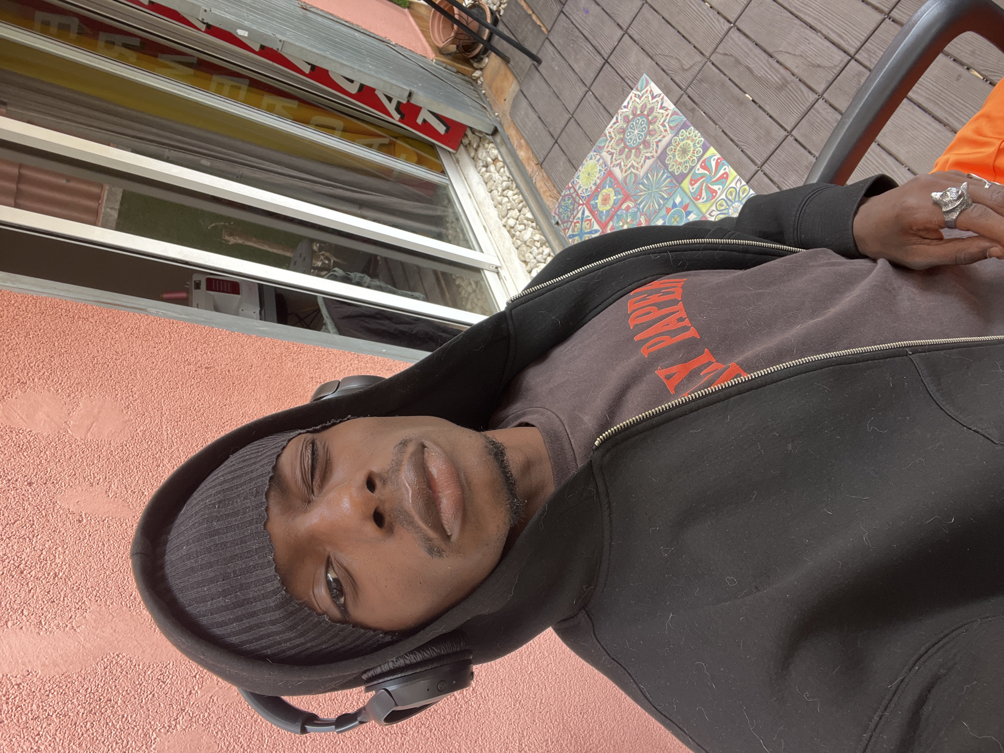 A medium close-up shot captures a person, likely male, with dark skin, wearing a black zippered hoodie, a dark brown t-shirt, and a dark beanie. Black over-ear headphones are around his neck. He has a short beard and mustache and is looking directly at the camera with a neutral expression, his head slightly tilted. His right hand is visible, resting near his torso, wearing a silver ring shaped like a wolf's head.
The scene depicts an outdoor setting, likely a patio or balcony in Barcelona, Spain. Behind the person's head is a textured, salmon-pink colored wall. To his right, the ground features a vibrant, multi-colored patterned tile, followed by wooden decking and a section of light-colored gravel. In the background, a building with windows is visible, topped by a red and white striped awning that has partial white text, possibly "SEVEN" or similar. Through the windows, greenery and a terracotta pot are seen, along with a white machine with pink accents in what appears to be an interior space. The red capital letters on the person's t-shirt appear to spell "EVERYDAY". The natural light suggests it is daytime.