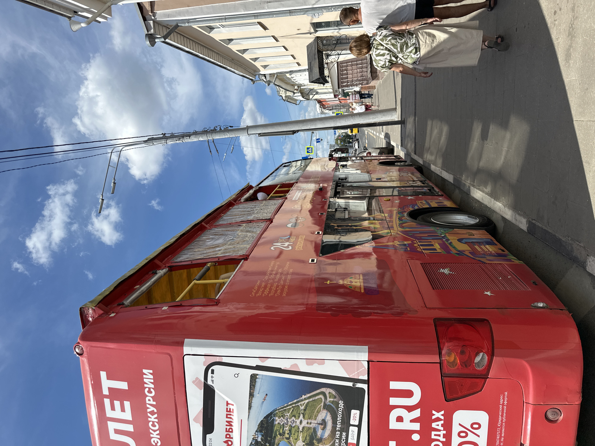 A bright red double-decker bus is parked on a street in a European-style city, likely in Russia given the Cyrillic text on the bus. The bus is adorned with colorful illustrations, possibly depicting local landmarks or cultural motifs. The words "ЛЕТ" and "ЭКСКУРСИИ" are visible on the side of the bus, indicating it is for summer tours or excursions. A portion of a smartphone screen is displayed on the bus, showing an image of a park with a body of water and a ship, alongside text that reads "ОРБИЛЕТ" and mentions discounts. The street is paved with stone, and buildings with light-colored facades and windows line the right side of the road. A utility pole with multiple wires stands near the bus. Two people, a woman and a man, are walking away from the camera on the sidewalk, with the woman wearing a patterned shirt and a long skirt, and the man in a t-shirt and shorts. The sky is blue with scattered white clouds. The overall impression is a sunny day in an urban environment. The location mentioned in the prompt, Putyatino (Semivragovskaya S/A), Russia, is a small village in the Ryazan Oblast, which is unlikely to feature a double-decker tour bus of this scale. Therefore, it is more plausible that the image depicts a tour bus in a larger Russian city.