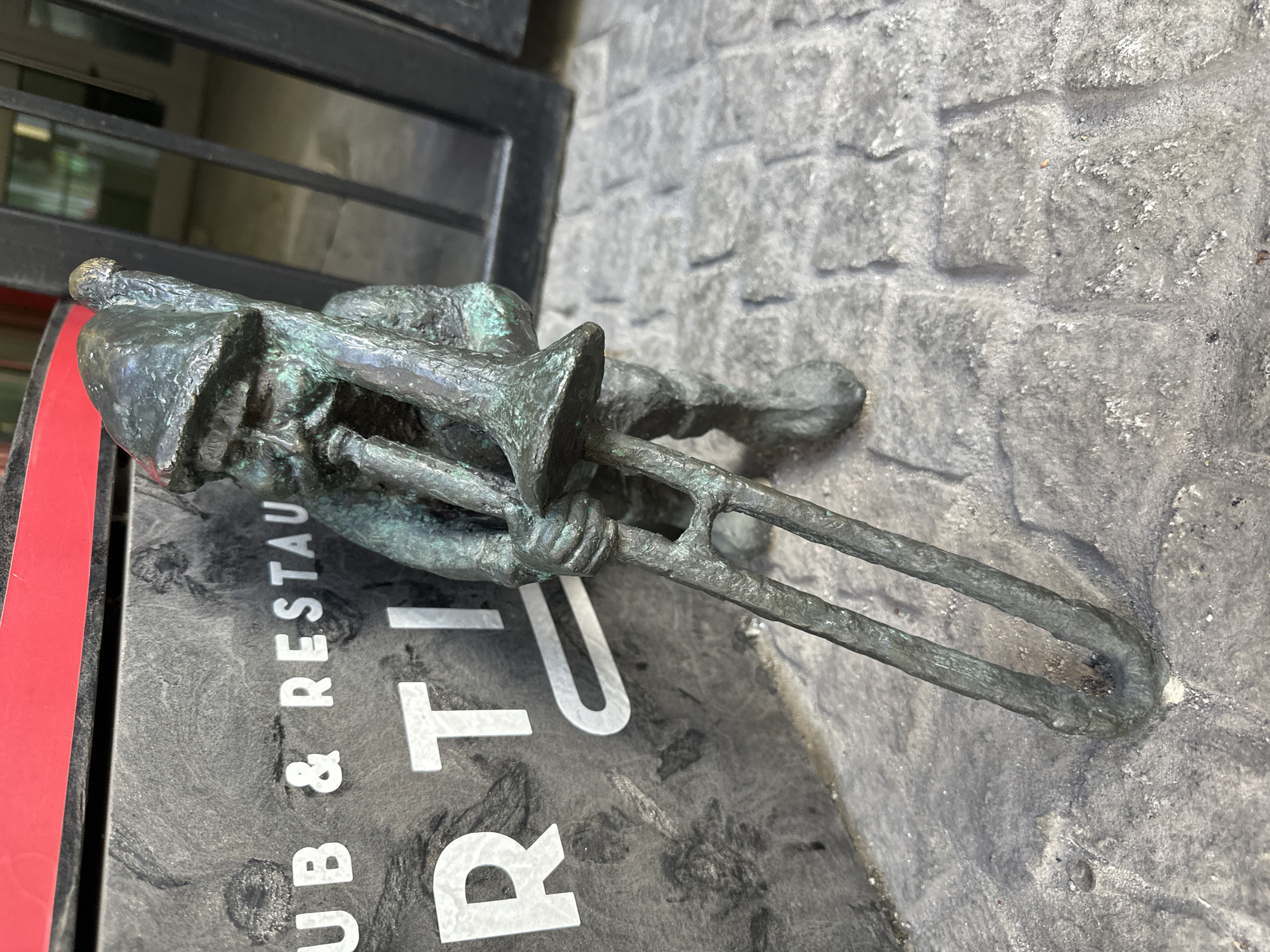 A small bronze statue of a gnome playing the trombone sits on a stone wall beside a sign for a pub and restaurant. The statue is weathered and appears to be outdoors. The image is taken at an angle, and the location is Wrocław, Poland.