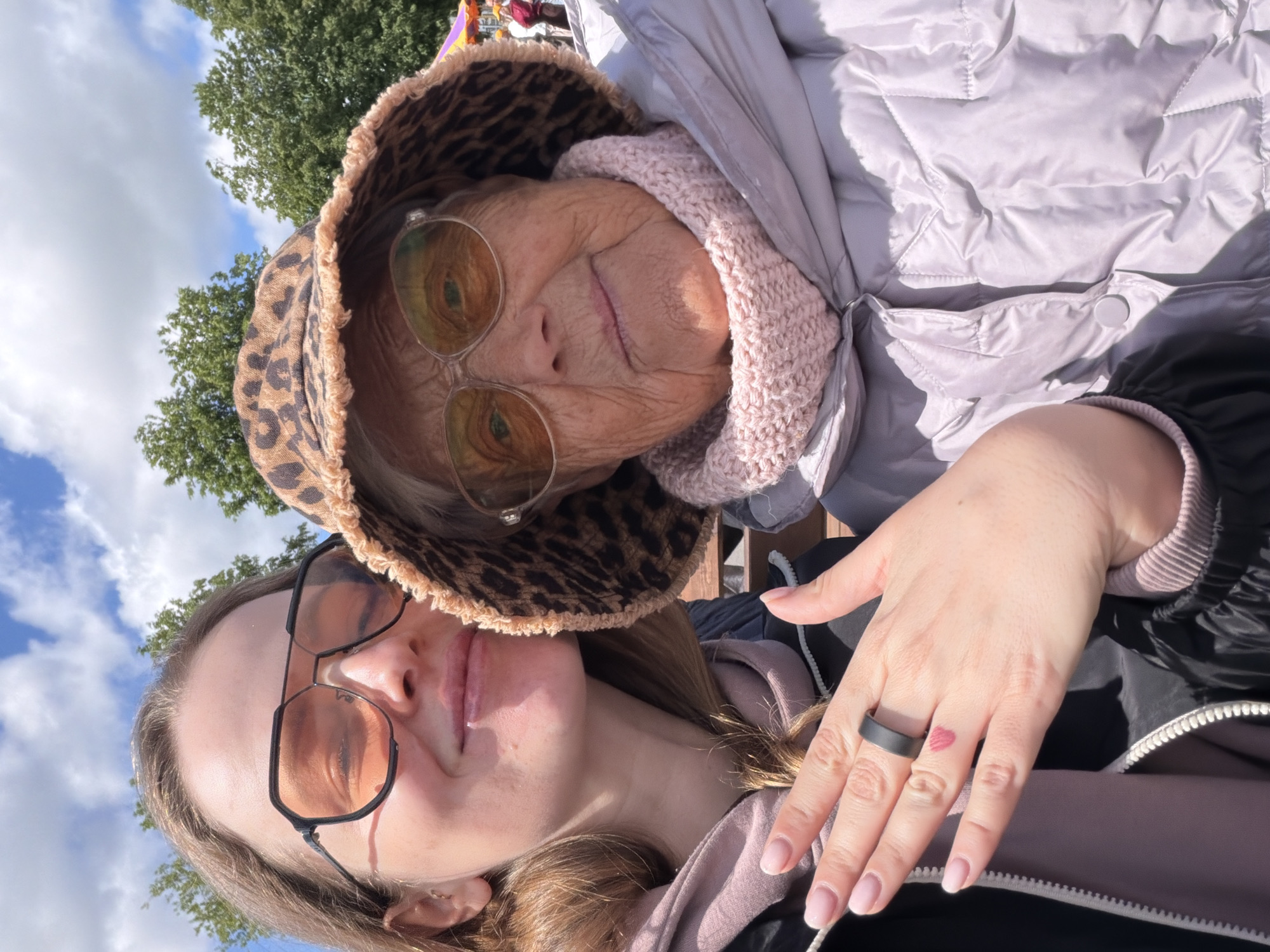 A low-angle selfie shows two people, an older woman and a younger one. The older woman is positioned at the top of the frame and slightly to the right. She wears a leopard-print bucket hat, glasses with reflective lenses, and a light pink knitted collar under a puffy silver jacket. The younger woman is positioned at the bottom of the frame and slightly to the left. She wears glasses with black frames and a pink hoodie under a black jacket. The younger woman's hand is visible on the right side of the frame, with a black ring on the ring finger and a small heart tattoo on the back of her hand. The background shows trees, a partly cloudy sky, and possibly other people in the distance. The location, Valozhyn, Belarus, is not discernible from the image itself.