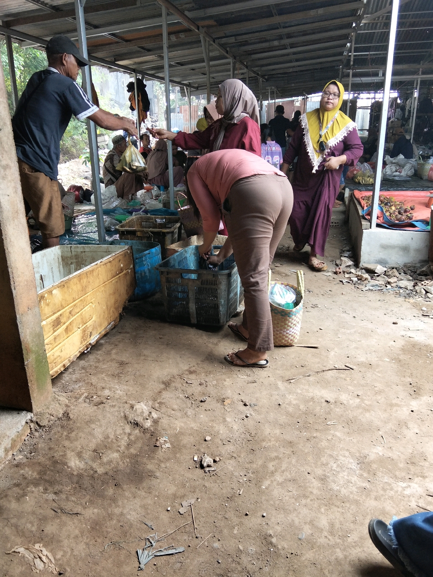 This image captures a scene at an outdoor market in Tirto Sari, Indonesia. In the foreground, a person wearing brown pants and a pink shirt is bent over, sorting through a blue plastic crate. Their feet are clad in black flip-flops. To the left, a large, weathered yellow plastic container is partially visible, alongside other plastic crates of various colors. In the mid-ground, a man wearing a dark blue t-shirt, brown pants, and a black cap is interacting with individuals seated behind a stall. He appears to be handing something in a plastic bag to a woman wearing a maroon headscarf and a maroon garment. Another woman in the background wears a yellow headscarf and a purple dress. The market environment is characterized by a corrugated metal roof supported by metal poles, with dirt flooring scattered with debris. Various items are visible in the background, including what appear to be produce and other market goods.