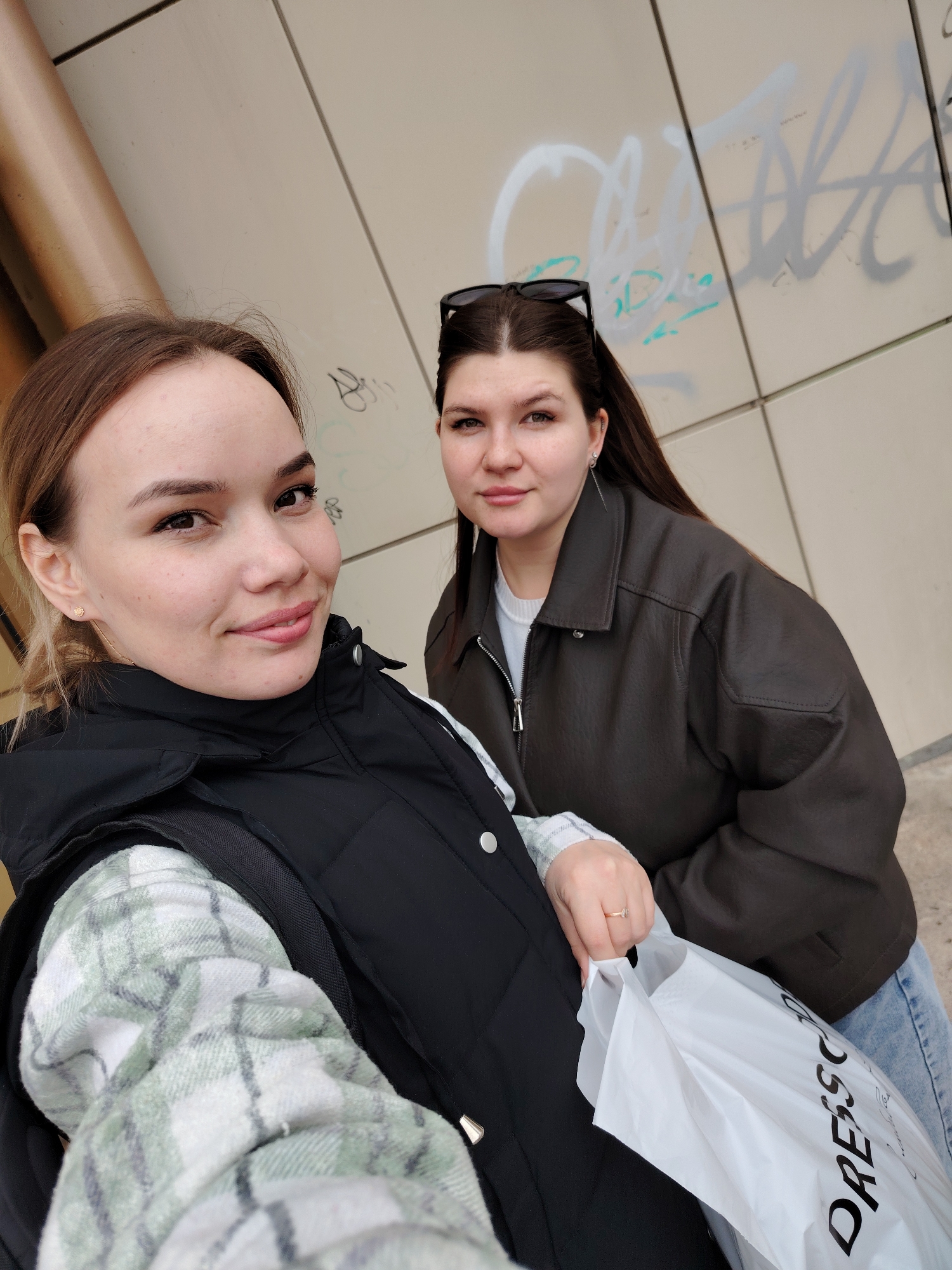 The image captures two female subjects, likely young adults, positioned outdoors in an unspecified area of Chaikovsky, Russia. The subject on the left, closer to the camera, has light brown hair and fair skin, with a light application of makeup, including eyeliner and lip color. She is wearing a black quilted puffer vest over a light green and white plaid long-sleeved shirt, with a backpack strap visible over her left shoulder. A small gold stud earring is present in her left ear. She is smiling and looking directly into the camera, which she appears to be holding for a selfie.
The subject on the right, positioned slightly behind and to the right of the first subject, has dark brown hair with black sunglasses resting on top. Her complexion is fair. She is wearing a dark brown leather-like jacket over a white zippered top, with light blue jeans visible below the waist. A silver chain earring is visible in her left ear, extending from a possible helix piercing. She maintains a neutral expression, looking directly towards the camera. Her right hand, adorned with a gold ring featuring a single clear stone on the ring finger, holds a white plastic shopping bag with the black text "DRESS CODE" prominently displayed.
The background consists of a light-colored, possibly beige or pale grey, paneled wall. Multiple layers of graffiti tags, primarily in light grey and pale green spray paint, are visible on the wall, particularly behind the right subject. In the upper left corner of the frame, a curved, light brown, metallic pipe or structural element is present. The lighting is even, suggesting an overcast day or shaded environment.