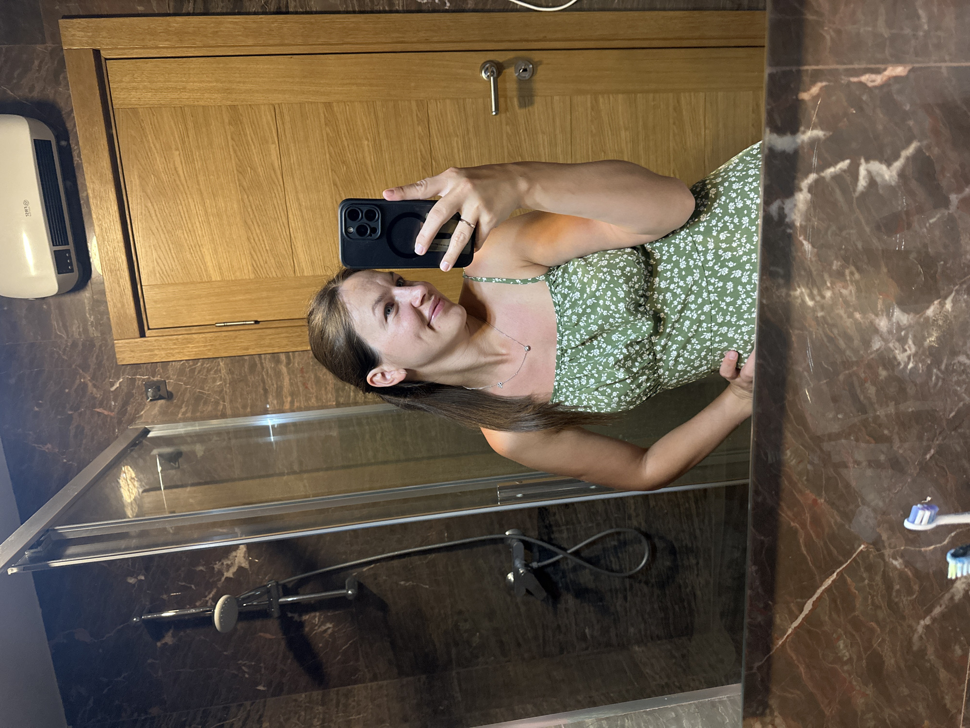 In a bathroom with dark marble countertops and walls, a woman with brown hair pulled back and a floral green dress poses for a mirror selfie. She holds a black smartphone in her right hand, with a silver ring on her left ring finger. The background shows a shower enclosure with a chrome showerhead and a wooden door with a silver handle. A white heater is mounted on the wall above the door. The countertop reflects two toothbrushes. The image was captured in Donji Pobori, Montenegro.