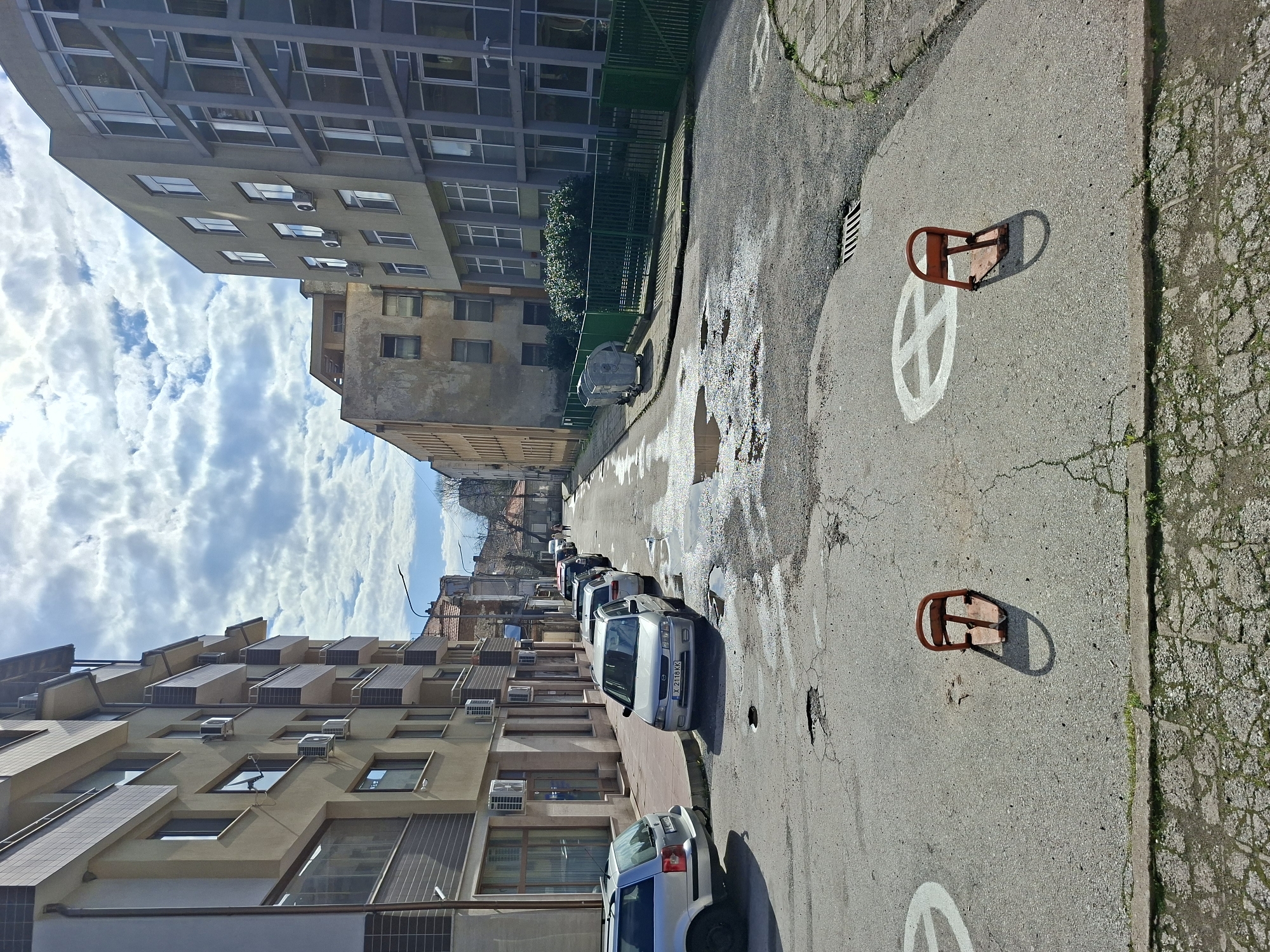 A street in Haskovo, Bulgaria, shows parked cars lining one side of the narrow road. The buildings along the street vary in height and architectural style, some modern and some older. The road surface is cracked and uneven, with patches of repair and standing water. White painted circles with an internal cross are spaced along the street. Two orange parking barriers are visible on the road. The sky above is partly cloudy.
