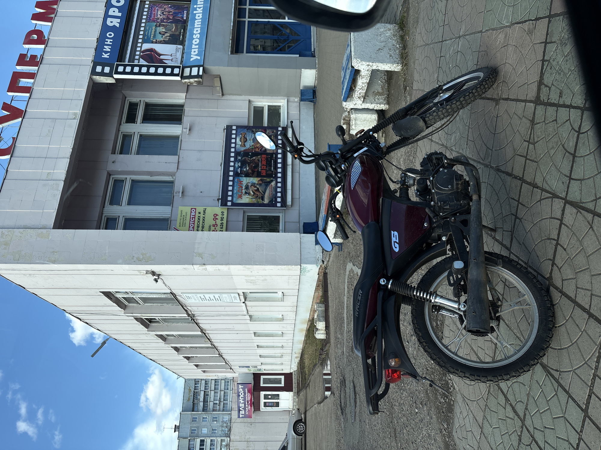 A maroon Racer GS motorcycle is parked on a patterned brick sidewalk in Ust-Ilimsk, Russia. The bike is positioned near a pale building identified by a large sign reading "КИНО ЯРС" (KINO YARS) and "СУПЕРМ". Movie posters are displayed on the building's facade. Adjacent to the movie theater, a sign for "ТЕЛЕПОРТ" (TELEPORT), a virtual reality club, is visible. Also near is a sign for "БЮРО ПО ЗАЩИТЕ" (BUREAU FOR PROTECTION) and other entities. An apartment building appears in the background under a cloudy sky. Part of a car is seen down the road. The image appears to be taken from the inside of a vehicle, as part of a car mirror is visible.