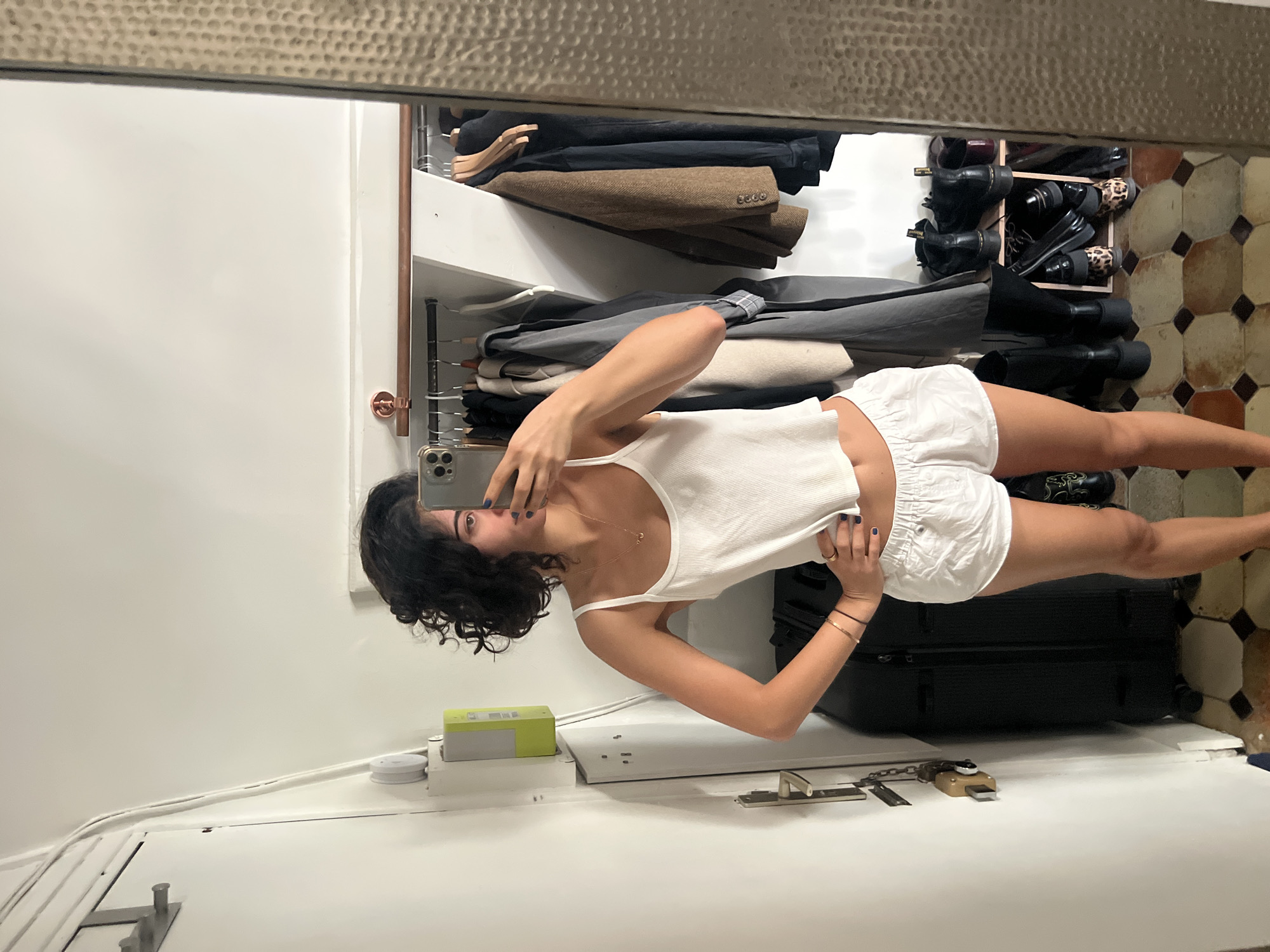 A person with dark, curly hair stands in front of a mirror, taking a selfie. They are wearing a white tank top and white shorts. In the background, a closet is visible with clothes hanging and shoes on shelves. The floor is tiled with a geometric pattern. The scene appears to be an indoor space, possibly a bedroom or dressing area. No specific location in Paris, France can be determined from the image.