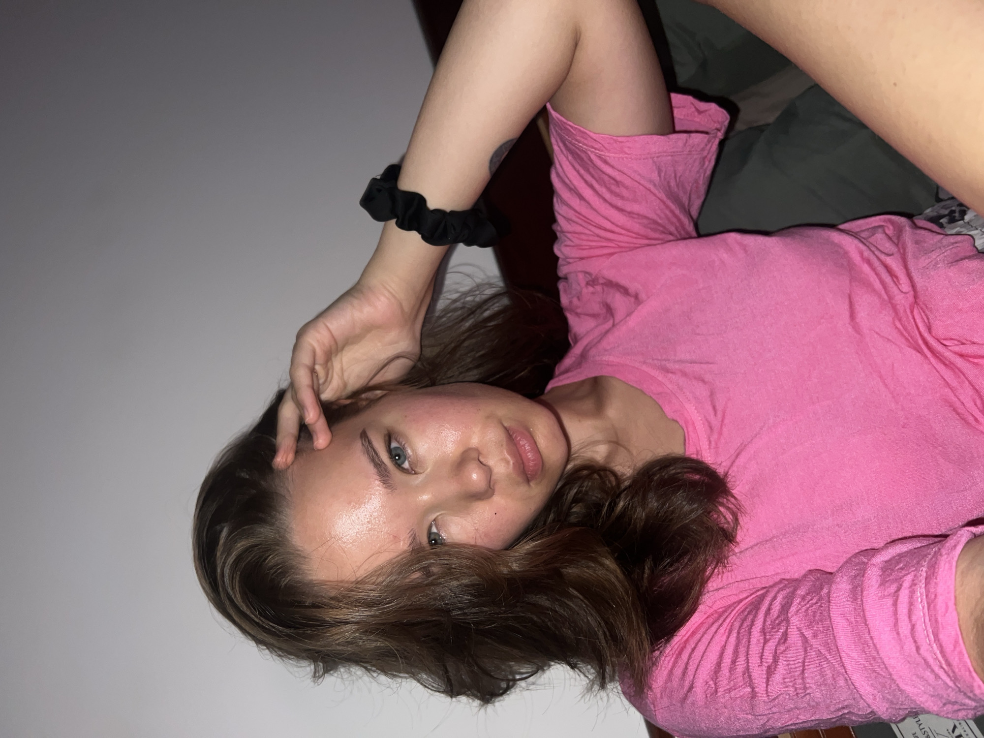 A woman is lying on a bed, wearing a pink t-shirt and a black scrunchie on her wrist. She has brown hair and blue eyes, and her hand is touching her forehead. The background consists of a white wall and a glimpse of bedding. The location of Nam Natural Heritage Area Trang, Vietnam, is not visible in the image.
