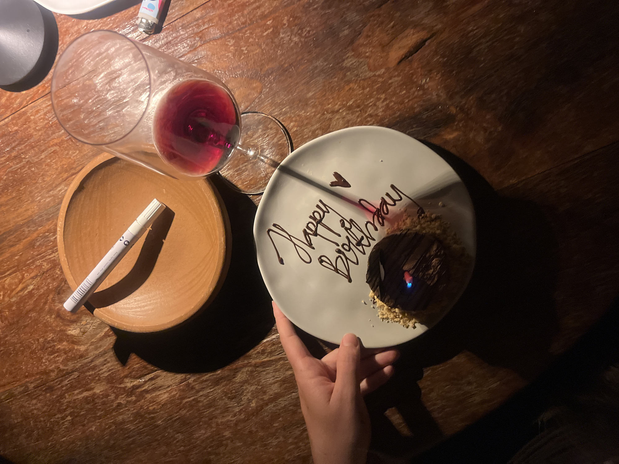 The scene presents a top-down view of a wooden table in Kuta Selatan, Indonesia, with various items placed on it. A half-full wine glass sits next to a small, light-brown wooden bowl containing a white marker. A white plate with chocolate writing of "Happy Birthday" encircles a dark chocolate dessert topped with what appears to be chopped nuts. A lit candle sits on top of the dessert. A hand is seen holding the edge of the plate. The lighting is subdued, casting shadows and highlighting the wood grain of the table.