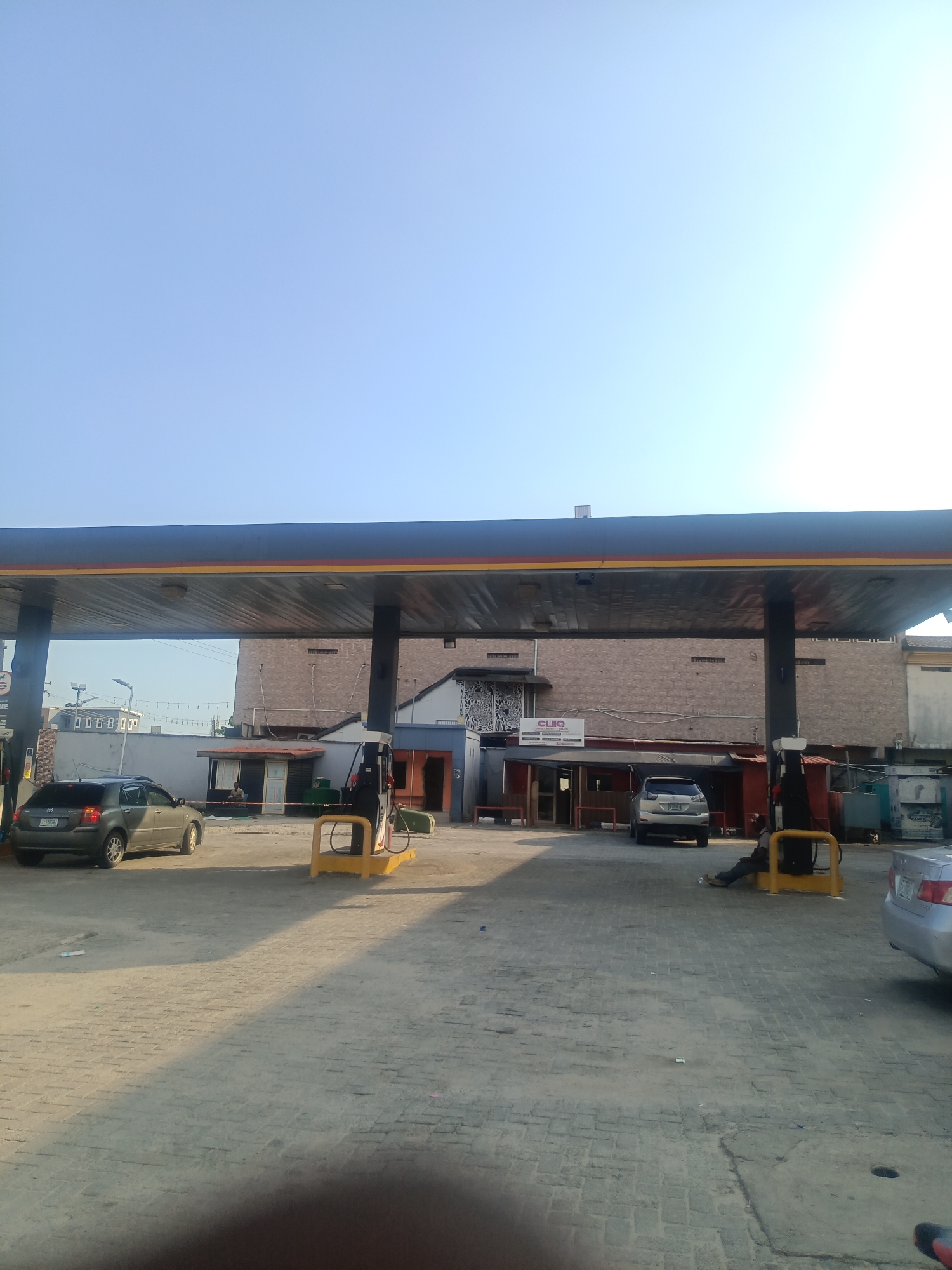 A gasoline station in Ikorodu, Nigeria, is depicted under a bright, clear sky. The station has a covered canopy supported by black pillars. A gray car is parked near a gas pump on the left, while a silver SUV is positioned at another pump on the right. A person is seated near the pump on the right, appearing to rest or wait. In the background, buildings with brick facades are visible, and there are other structures that suggest commercial activity. The ground is paved with interlocking bricks, and shadows are cast across the scene, indicating it's daytime.