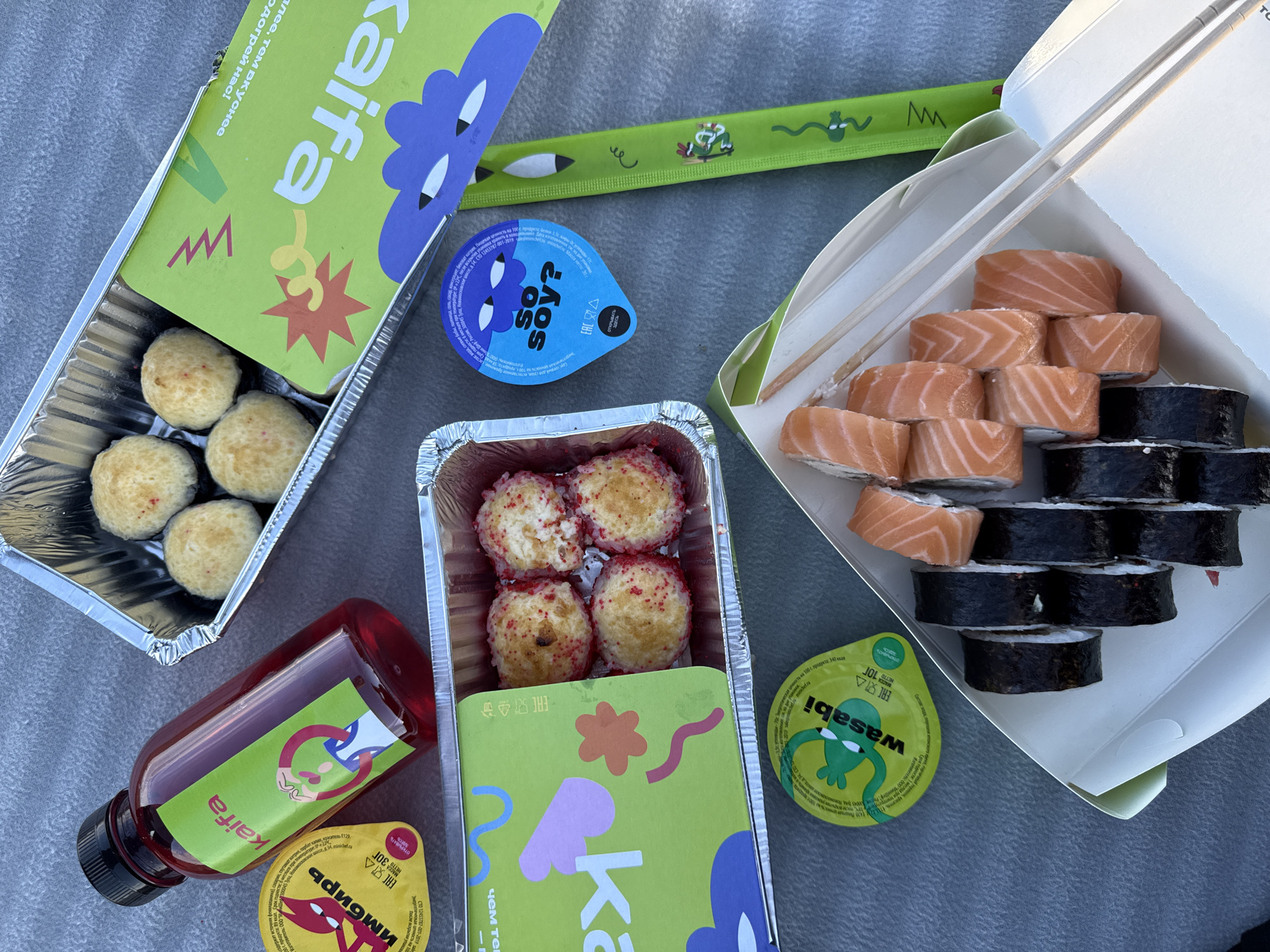 A picnic scene is laid out on a grey, textured surface, possibly a blanket or tablecloth. In the center of the frame, a white cardboard box contains a serving of sushi rolls. The rolls are cut into pieces, with the salmon-topped ones visible on the right and darker, nori-wrapped rolls on the left. Two wooden chopsticks are placed on top of the sushi. To the right of the sushi box, a green paper wrapper for chopsticks is visible, featuring a playful depiction of a frog.
Above the sushi box, two foil containers hold various food items. The left container holds several round, breaded balls, some of which are sprinkled with red roe. The right container appears to be empty.
To the left of the foil containers, a red liquid is contained within a clear bottle with a black cap. A green label on the bottle features cartoonish graphics and the word "kaifa" in pink lettering. Next to the bottle, a small yellow container with a red devil-like cartoon and text is also visible.
Below the foil containers, a small blue container with a cartoon face and the text "so soy?" is present. Further down, a green squeeze packet labeled "wasabi" is depicted. The overall impression is of a takeaway meal intended for outdoor consumption. The reference to Chaikovsky, Russia, is not discernible from the visual information provided in the image.