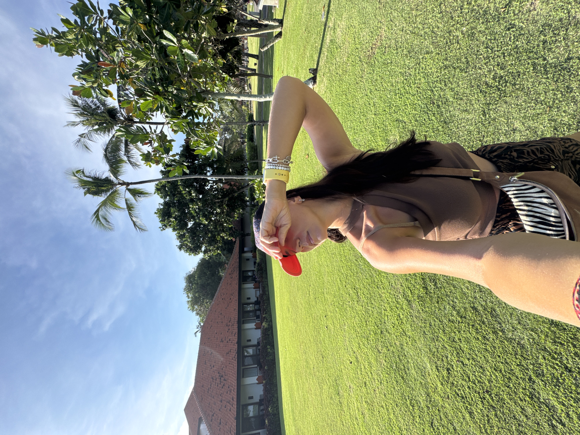 A selfie depicts a woman on green grass in Kuta Selatan, Indonesia. She wears a brown top, a red cap, and bracelets. A building with a red roof is visible, as are trees and a blue sky.