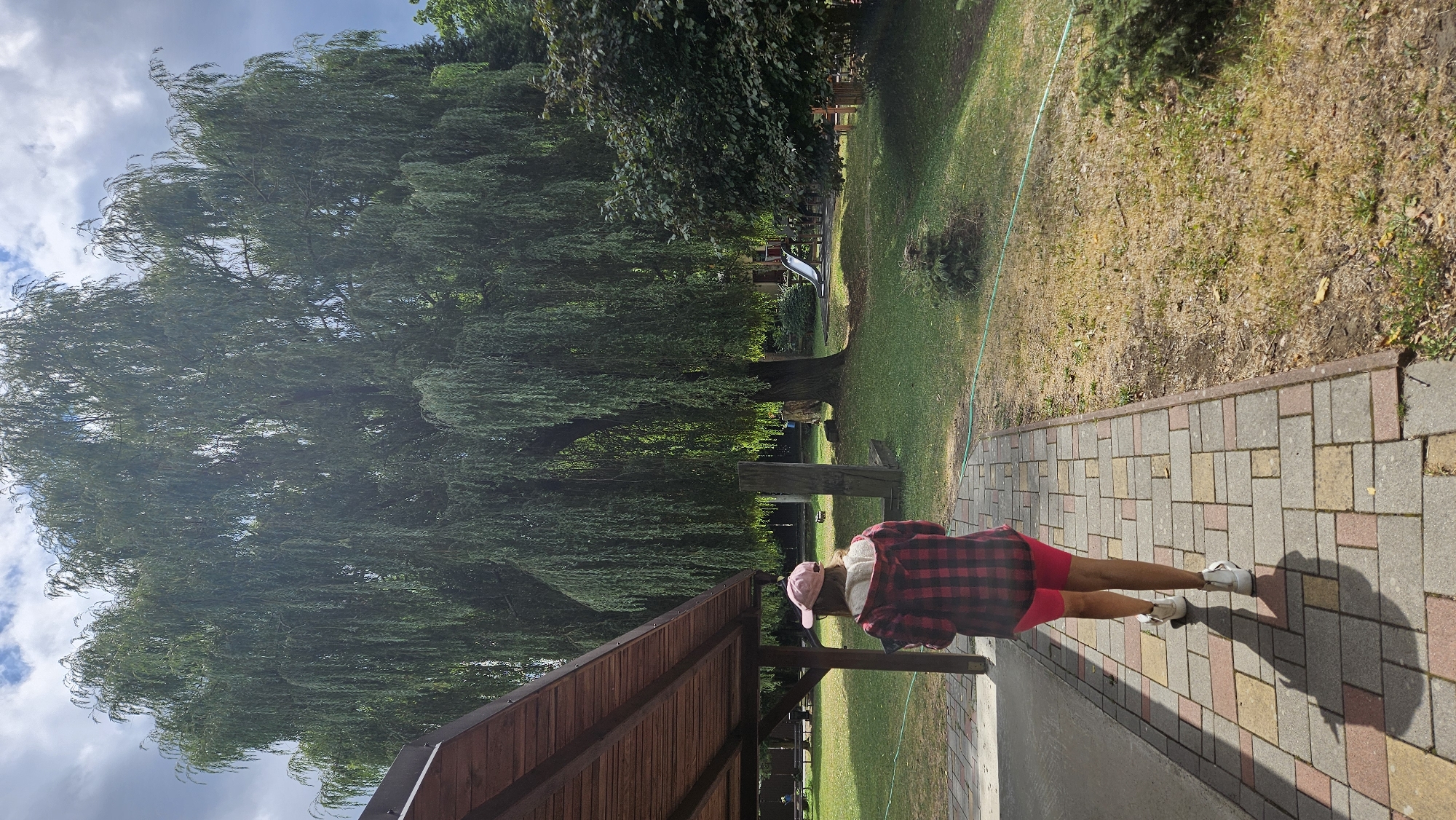 A person with their back to the camera, wearing a red and black checkered shirt, pink shorts, a pink cap, and white sneakers, walks away from a covered seating area on a paved walkway. The walkway is constructed with rectangular bricks in shades of red, yellow, and grey. The person is casting a shadow on the walkway. The background features a large weeping willow tree with its branches hanging low, a grassy area, a small evergreen tree, and what appears to be a playground with a slide. The scene is set outdoors in what could be a park or a recreational area. The image does not provide specific information about the location in the city of Prešov, Slovakia.
