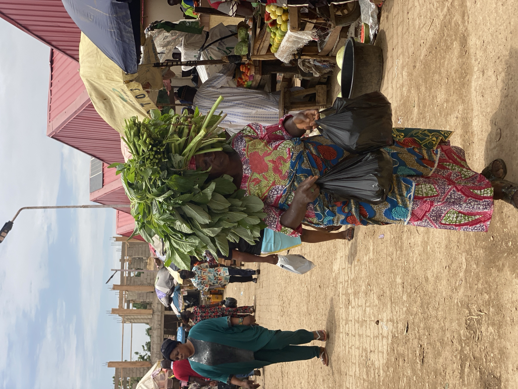 The image depicts a bustling marketplace in Kafanchan, Nigeria. In the foreground, a woman in a vibrant, patterned dress carries a large bundle of leafy green vegetables on her head, along with two black plastic bags held in her hands. The marketplace is crowded, with people walking around and vendors selling goods. The ground appears to be dirt and compacted earth. There's a building under construction in the background, along with other buildings with red roofs. The sky is partly cloudy. A variety of merchandise, including fruits and vegetables, can be seen at the vendors' stalls.