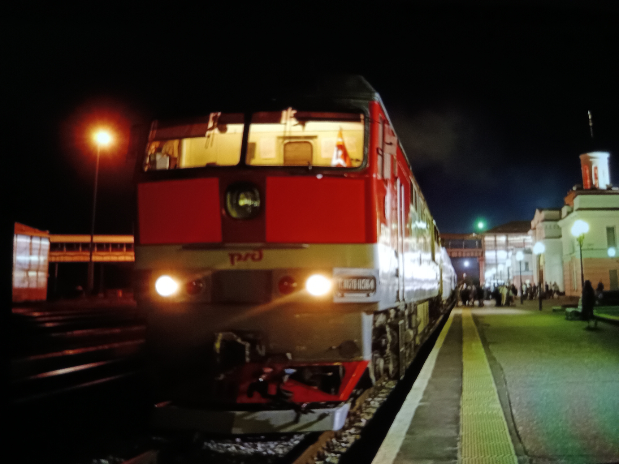 The image captures a night scene at a train station, likely in Košice, Slovakia, given the context. A red and gray train is prominently featured, stopped at the platform. The train's headlights are illuminated, casting a bright glow. The station platform is visible, with yellow tactile paving indicating the edge. In the background, a building with architectural details, possibly a station building, is illuminated, and several people are gathered on the platform. The overall atmosphere is that of a functioning train station at night.