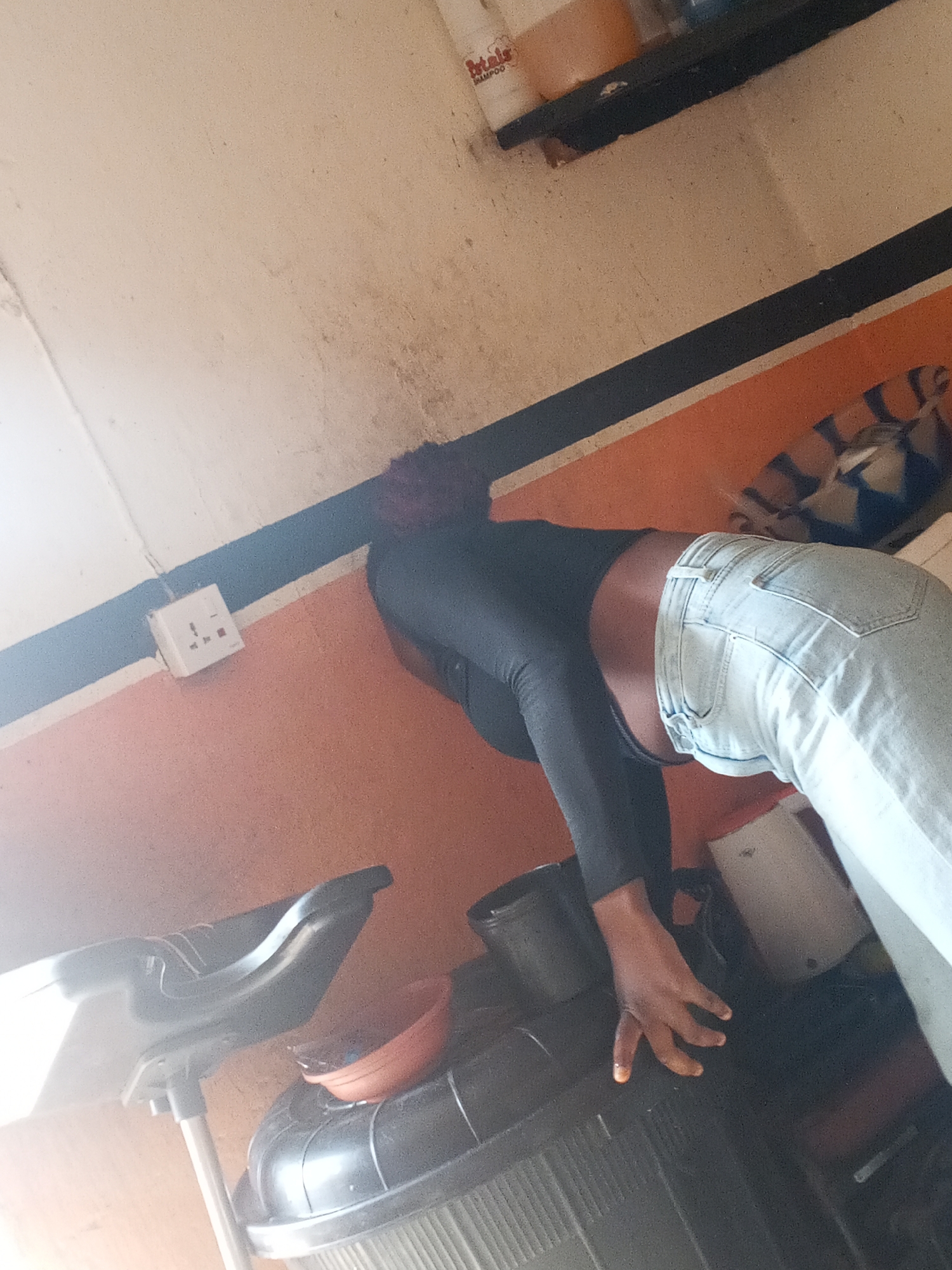 A woman wearing a black long-sleeved shirt and light blue jeans is leaning forward, her hand resting on a black plastic object. The background consists of a wall with alternating bands of white and orange paint, separated by a black stripe. A white electrical outlet is visible on the wall. A shelf containing bottles is present in the upper portion of the image. Other indiscernible objects surround the woman. The scene is located in Gwong, Nigeria.