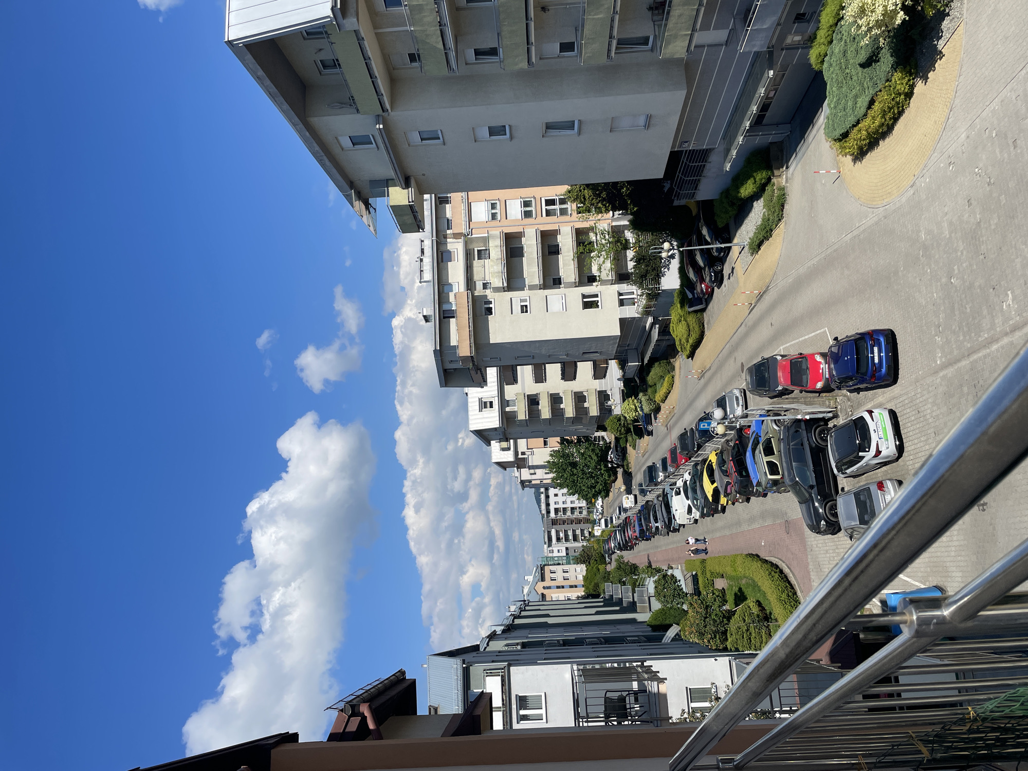 The image shows an outdoor urban environment. The scene is observed from an elevated position, likely a balcony or upper floor of a building, as indicated by a metallic railing in the foreground. A row of multi-story buildings lines the street. A variety of parked cars occupy the street, showcasing colors like blue, red, yellow, and white. There are sidewalks with manicured green hedges along the sides. The sky above is bright blue, with puffy white clouds scattered throughout. People are not distinctly visible in the scene. The general location is Unknown, Unknown.