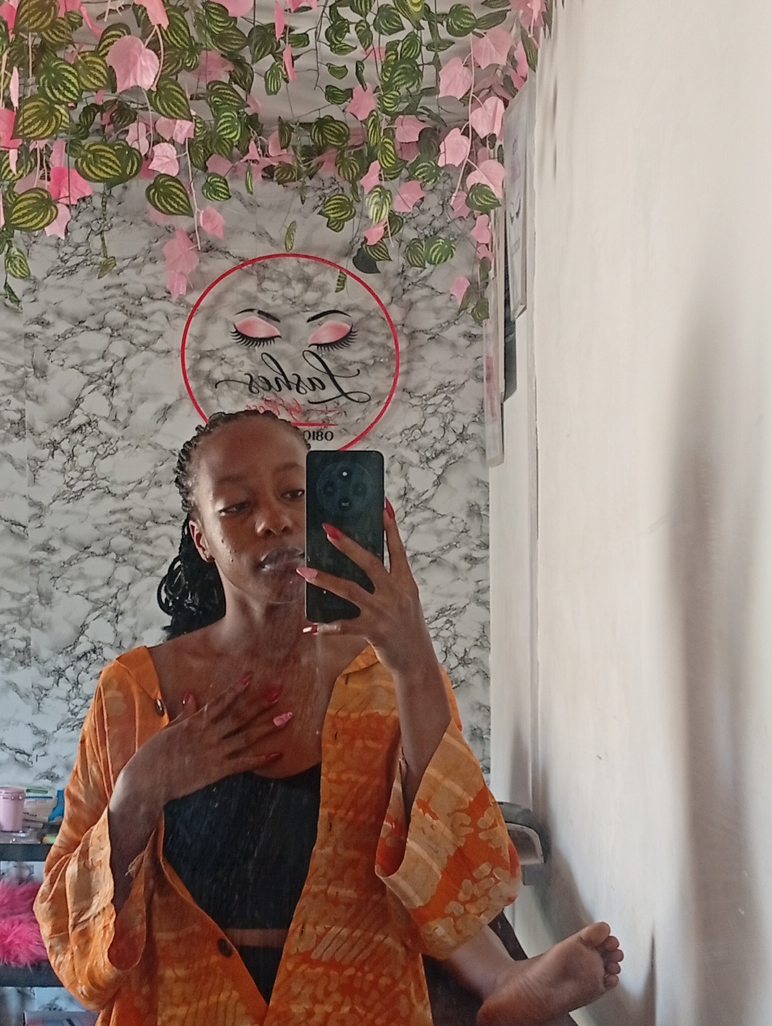 A person is taking a selfie in front of a mirror. The person is wearing a black top and an orange patterned shirt. The background behind the person features a white marble-patterned wall with decorative artificial vines and a circular sign with a stylized eye logo. The scene appears to be indoors, possibly in a salon or beauty-related business located in Narayi, Nigeria. A portion of a bare foot is visible in the lower right corner.