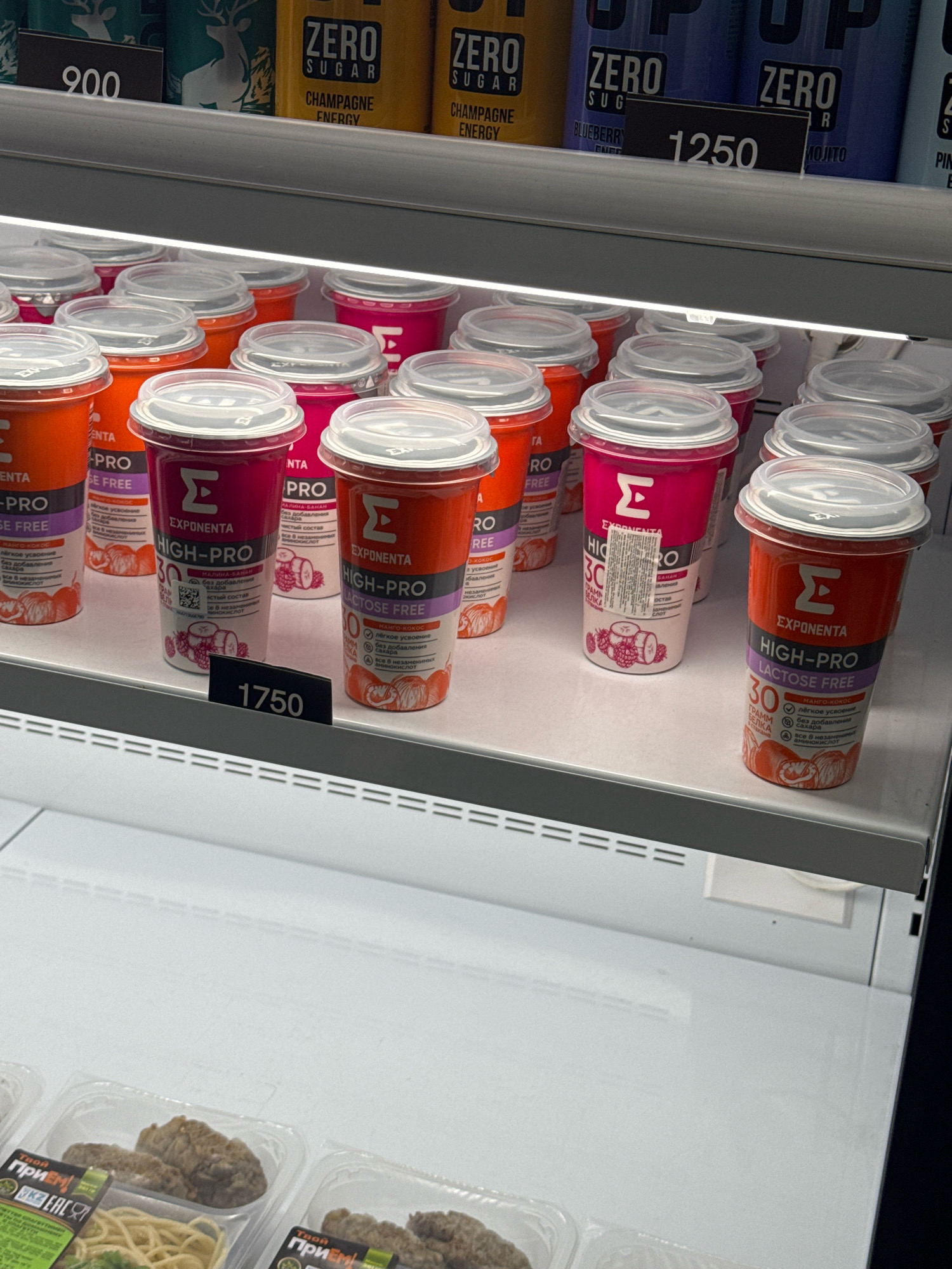 A close-up shot captures a refrigerated display case filled with several rows of drinks in plastic cups. The drinks appear to be some kind of protein or energy beverage, with branding visible on the cups. Many of the cups have clear plastic lids. In the foreground, a black price tag displays the number "1750". Above the display, a shelf holds cans of drinks with labels indicating "ZERO SUGAR" and various flavors like "CHAMPAGNE ENERGY", "BLUEBERRY", and "MOJITO". Another price tag in the background shows "1250". The lighting in the display case is bright, illuminating the products. The overall environment suggests a retail setting, likely a convenience store or supermarket. The presence of specific brands and pricing suggests this scene is located in Almaty, Kazakhstan, given the implied context of the city.