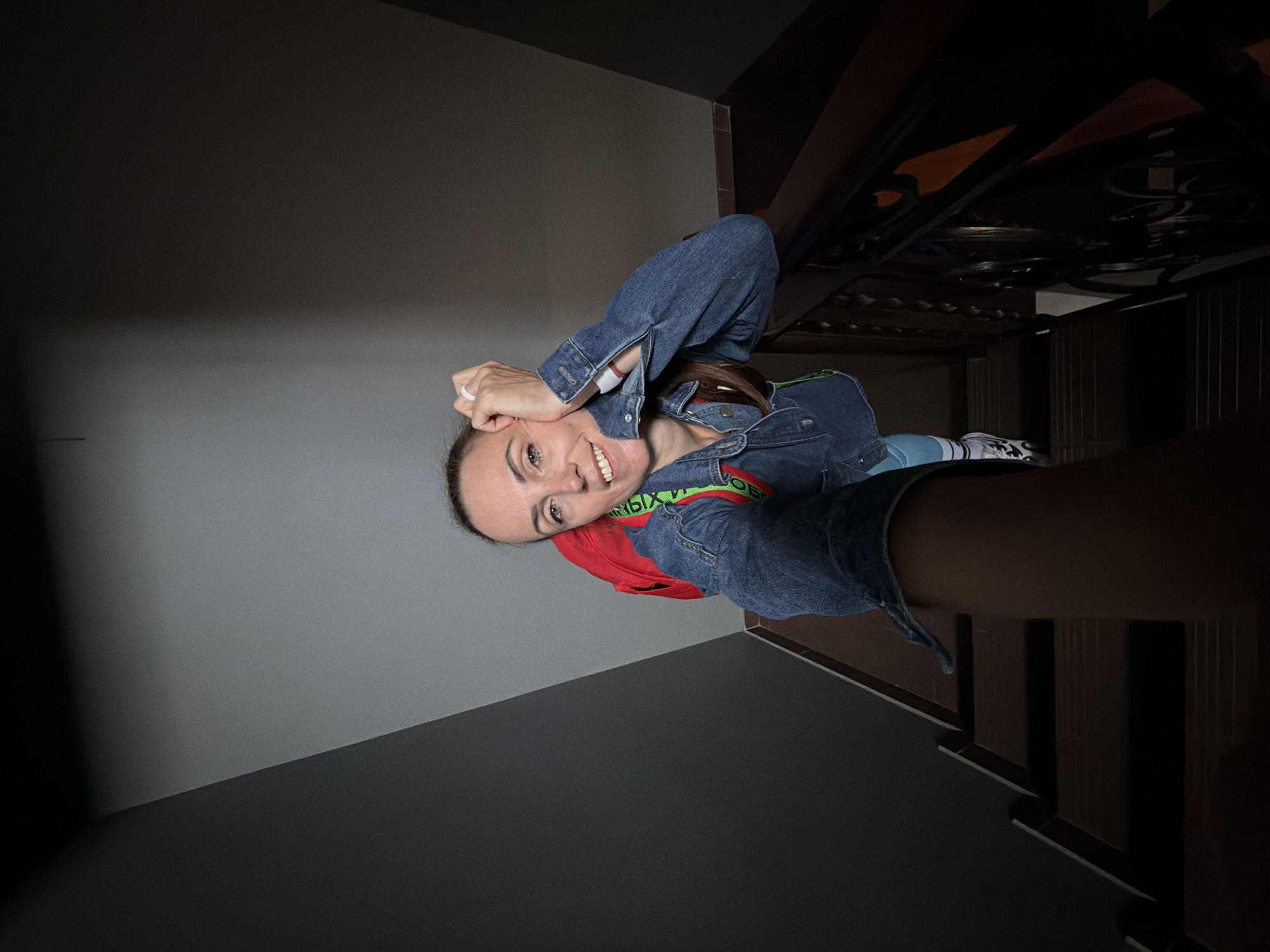A woman is posing for a selfie on a staircase. She is wearing a denim jacket, a red backpack, and light blue pants. She has her right arm resting on her head and is smiling at the camera. The staircase is made of dark wood with a metal railing. The walls are painted gray and dark gray. The image does not provide any information about the location being Birobidzhan, Russia.
