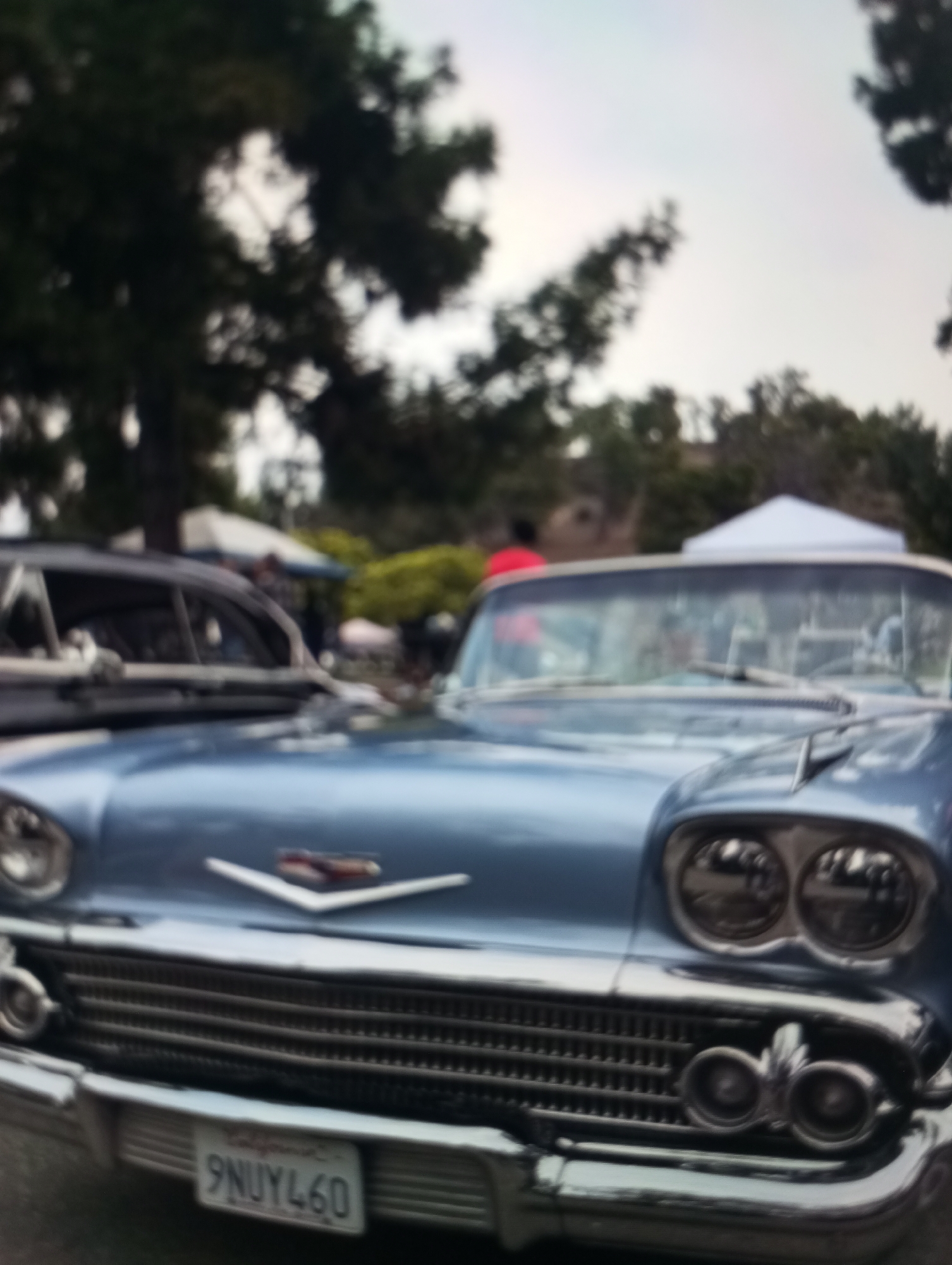 The image shows a close-up of a light blue vintage car, likely a convertible, parked outdoors. The car's front grille, headlights, and license plate are clearly visible. The license plate reads "9NUYL60". The background is blurred, showing trees, a white tent, and what appears to be other vehicles and people, suggesting a car show or outdoor event. The overall setting is outdoors with natural light. There is no visible indication of the car's location being in the city of Zahorna, Moldova.