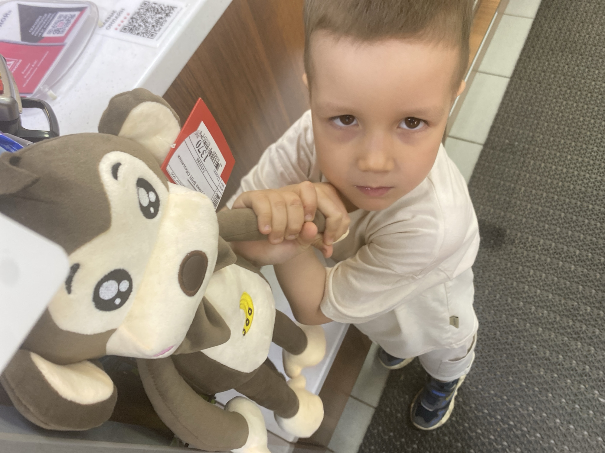 A young boy is standing in what appears to be a store in Chaikovsky, Russia. He is wearing a light-colored t-shirt, light-colored pants, and blue and black sneakers. He is holding a plush monkey toy with a price tag attached. The location has a white countertop, a wooden surface, and a grey tiled floor. There is a QR code and what looks like a promotional card on the countertop.