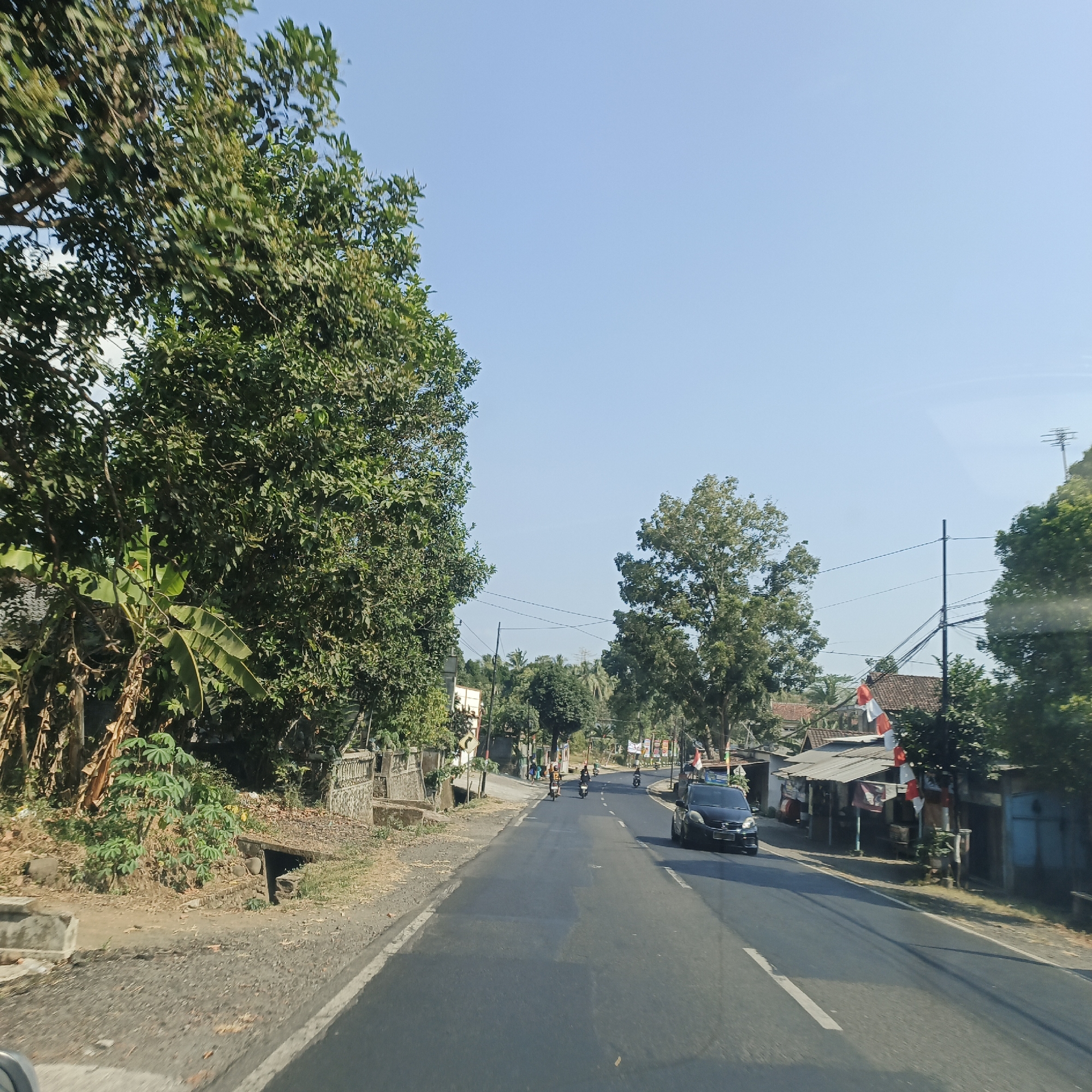 The image depicts a sunny day along a paved road in the city of Kebon Gunung, Indonesia. Lush green trees line the left side of the road, with some vegetation and a drainage ditch visible near the edge. The road itself is asphalt, marked with white lines. Several motorcycles and a black car are present on the road, with a few individuals riding the motorcycles. On the right side of the road, there are houses and small shops, some adorned with red and white flags, suggesting a local celebration or national holiday. Utility poles and wires are visible throughout the scene. The sky is clear and blue.