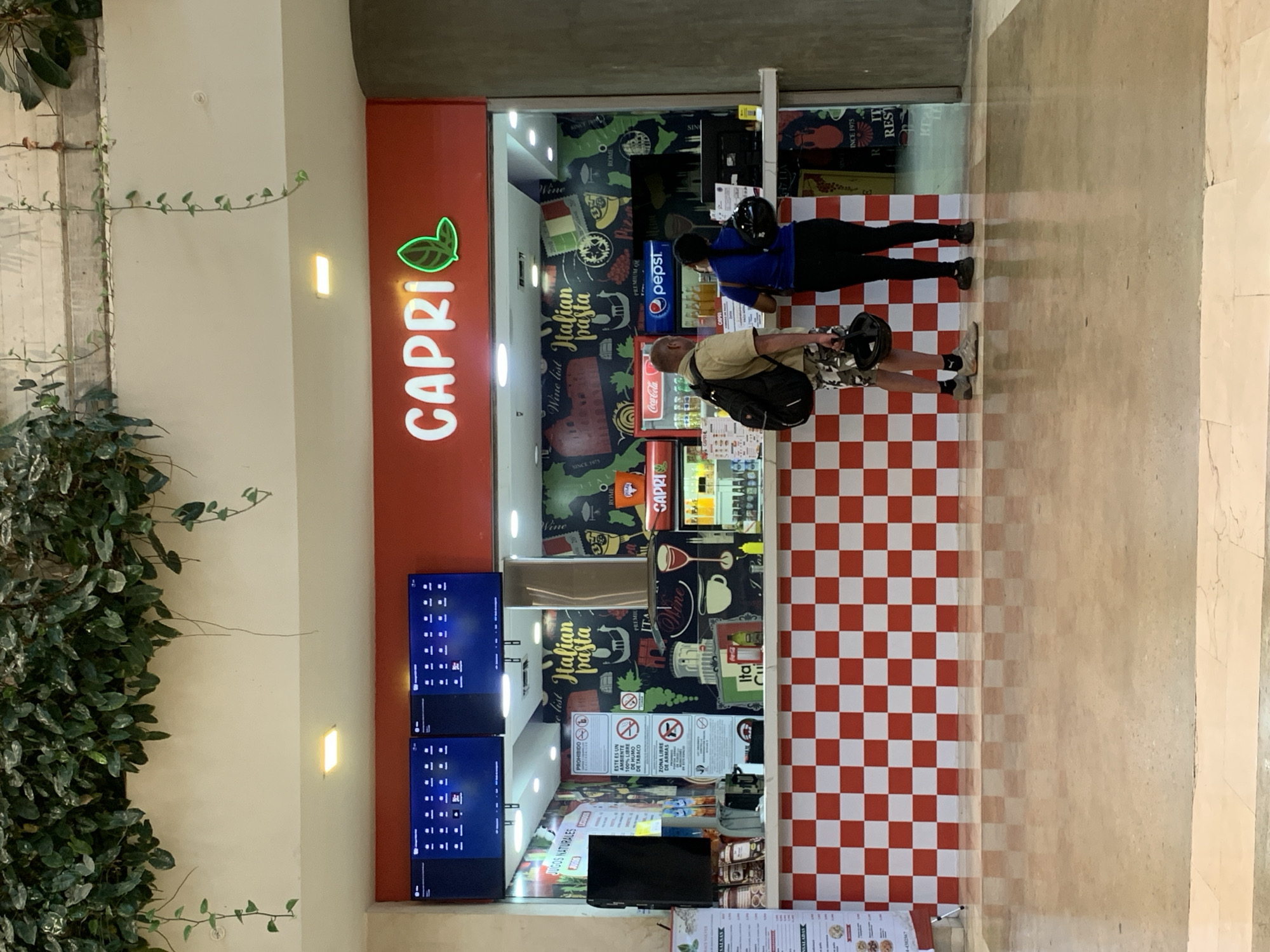 The image depicts an indoor scene at a food stall named "CAPRI Italian pasta" in Pampatar, Venezuela. The stall's facade is red, featuring the "CAPRI" logo in white letters with a green leaf design. Inside, the walls are adorned with Italian-themed graphics, including images of Italy, the Leaning Tower of Pisa, pasta, and wine glasses. The lower section of the stall and the adjacent floor space are covered in a distinctive red and white checkered pattern.
Two individuals are visible: a man wearing a tan shirt and a backpack, holding a drink, and a woman in a blue shirt carrying a black helmet. They appear to be customers at the counter, possibly placing an order or awaiting service. Behind them, drink refrigerators stocked with Coca-Cola and Pepsi are visible.
Several signs and texts are present. The stall advertises "Italian pasta" and "SINCE 1975." Other visible Spanish text includes "JUGOS NATURALES" (Natural Juices), "ESTE ES UN AMBIENTE 100% LIBRE DE HUMO DE TABACO" (This is a 100% Tobacco Smoke-Free Environment), and "ZONA LIBRE DE ARMAS" (Gun-Free Zone). Menu boards and additional text like "Wine list" and "ROME" are also discernible. The area is brightly lit, and green foliage from a plant is visible along the left side of the frame, suggesting an indoor garden setting nearby.