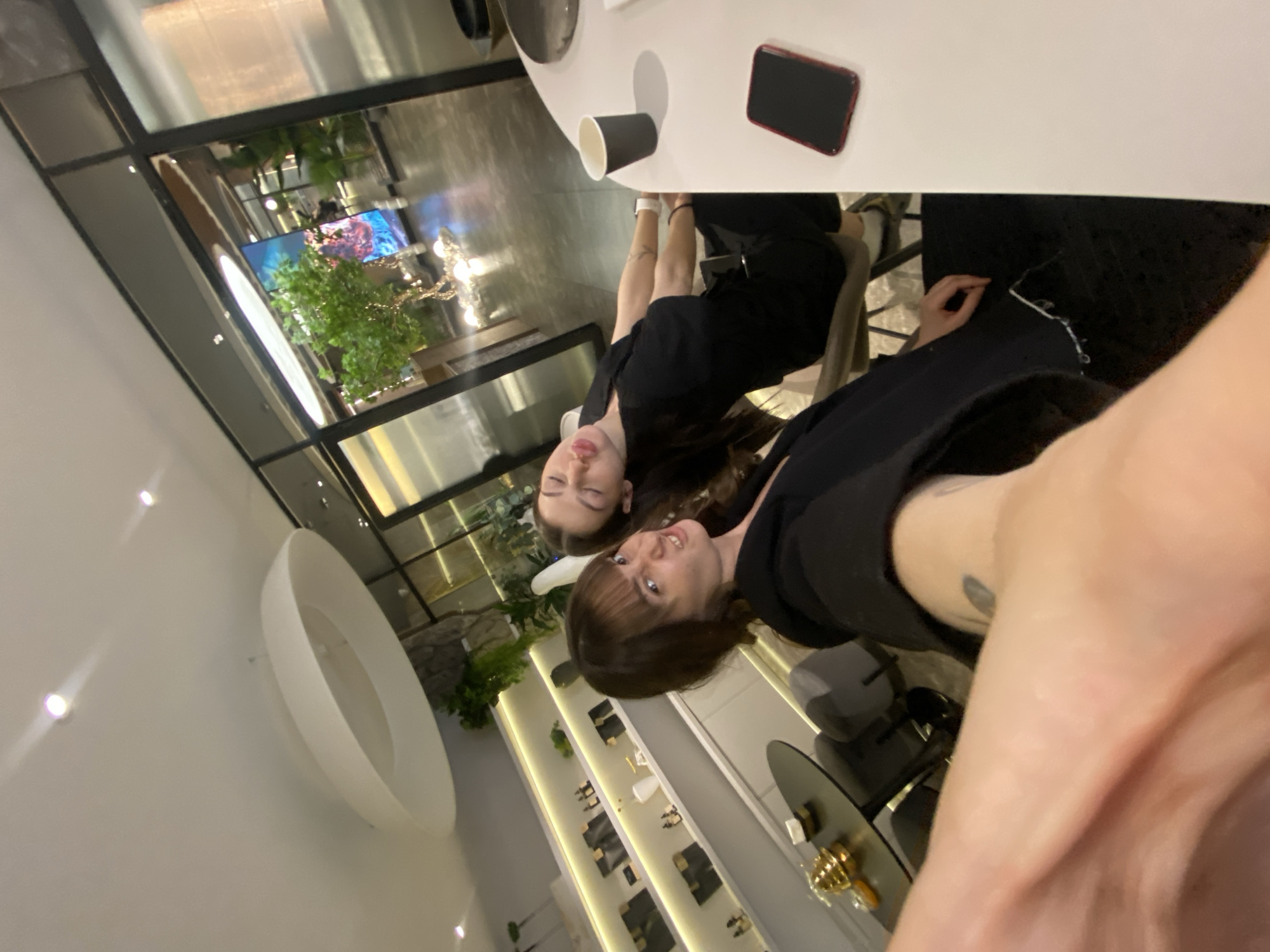 A selfie taken in a modern interior setting. Two people, likely female, are visible at a table. Both individuals are wearing black clothing. On the table are a black cup and a red smartphone. The room has a contemporary design, with white walls, track lighting, and decorative elements, including a large oval wall-mounted bowl. Outside the glass windows, a tree and decorative display is visible. The image appears to be taken indoors, during the day, at a commercial establishment. The presence of display cases suggests a shop or store. The location is unspecified.
