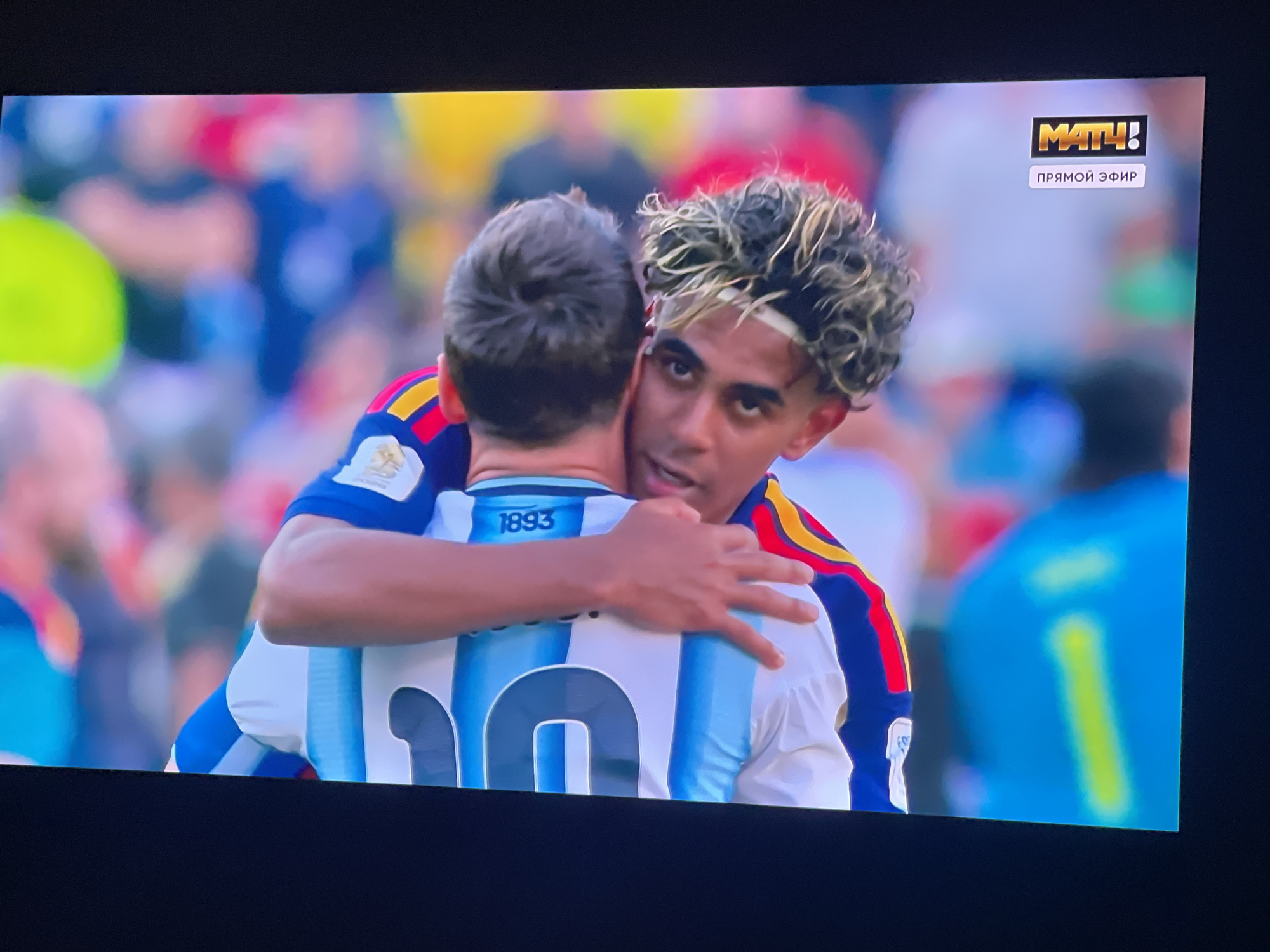 The image shows a screen displaying a football match. Two male football players are embracing, with one player's arms around the other's shoulders. The player on the left is wearing a blue and white striped jersey with the number "10" visible. The player on the right has curly, blonde-highlighted hair and is wearing a jersey with red and yellow stripes on the sleeves. In the upper right corner of the screen, there is a logo that reads "МАТЧ!" and below it, text that says "ПРЯМОЙ ЭФИР". The background of the screen is blurred, showing spectators and the stadium environment. The overall scene suggests a moment of camaraderie or celebration during a football game broadcast on a screen. The specific location within the city of Stary Oskol, Russia, cannot be determined from the image itself.