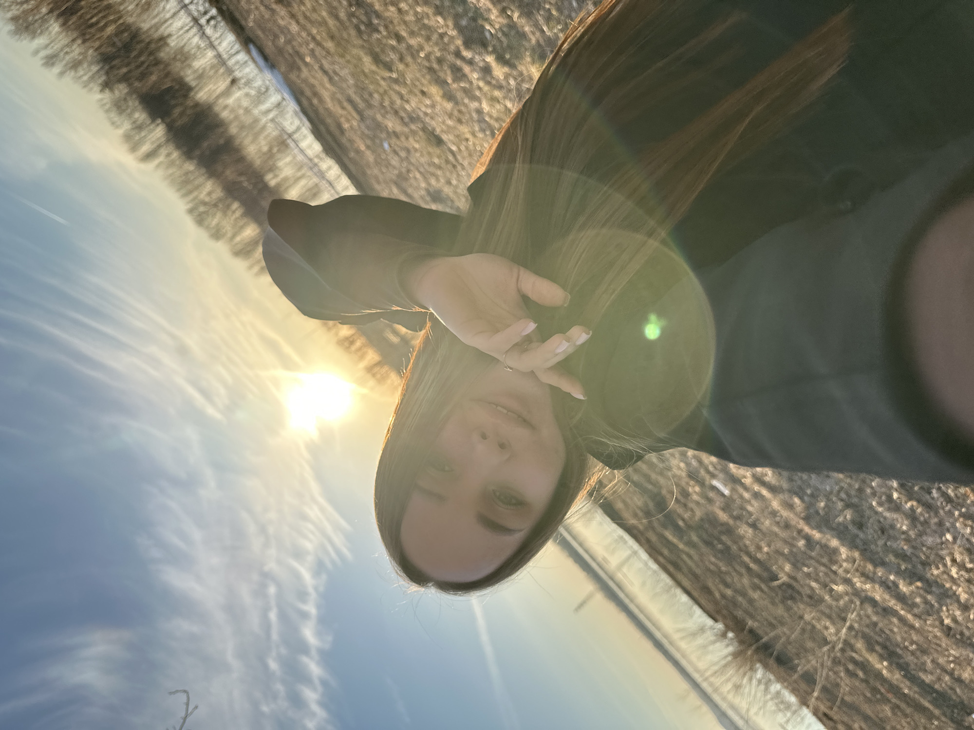A young woman is captured in a sun-drenched outdoor portrait, likely a selfie, in Chaikovsky, Russia. The image is taken during the golden hour, with the sun positioned behind and to the upper left of the subject, creating a prominent green and circular lens flare that washes over her hair and face. She has long, straight brown hair and light-colored eyes, gazing directly at the viewer with a neutral, slightly smiling expression. Her right hand is raised, partially obscuring the side of her face, revealing manicured nails painted in a light pink hue and a silver-colored ring on her ring finger. She wears a dark top or jacket.
The setting appears to be an open field or rural area. The background features dry, light brown ground or sparse vegetation, indicative of late autumn or early spring. A distant line of bare deciduous trees runs horizontally across the mid-ground, suggesting a natural boundary. The sky above is a clear blue with scattered delicate, wispy white clouds, and a faint airplane contrail can be seen in the upper left. The overall mood is warm and serene, highlighting the natural beauty of the setting and the soft, golden light of the declining sun. No visible text is present.
