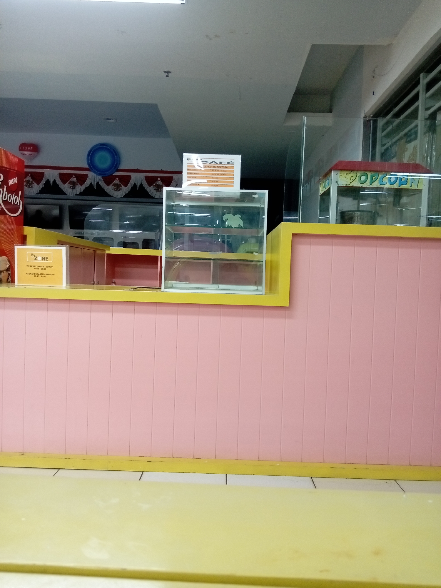 The image depicts an indoor scene featuring a concession stand with a pink paneled wall and a yellow countertop. On the countertop, there is a yellow sign with the text "play ZONE" and operating hours. Behind the countertop, glass display cases and a popcorn machine are visible. The background shows a ceiling with lighting fixtures and a glimpse of outdoor elements through windows. Based on the text on the signs and the overall aesthetic, this scene is likely in a commercial establishment within Batam, Indonesia. There are no people or actions depicted in the image.