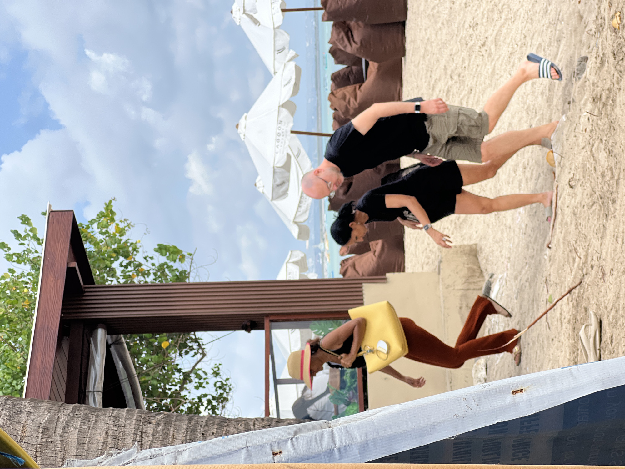 In Kuta, Indonesia, three individuals walk on a sandy beach. A woman wearing a straw hat with a pink band and a black tank top carries a yellow tote bag. She is walking to the left. Behind her, a woman in a black t-shirt and black shorts is walking to the left. A man wearing a black t-shirt and khaki shorts is walking to the right. In the background, there are beanbag chairs and white umbrellas. The sky is cloudy.