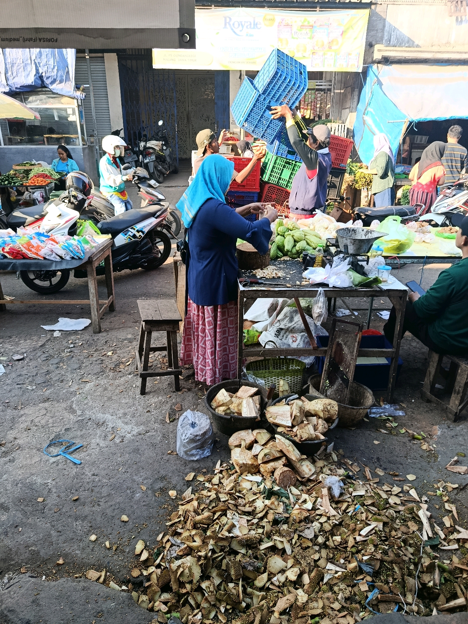 A street market scene is captured in Gondanglegi, Indonesia, with several individuals engaged in various activities amidst a cluttered environment. In the foreground, a significant pile of durian husks and waste is scattered across the concrete ground, indicating recent preparation or disposal of the fruit. Several baskets and containers are visible, some filled with durian chunks and others empty.
A woman wearing a blue hijab and a long, patterned skirt stands near a table laden with produce, including what appear to be chayotes and other vegetables. She is actively involved in sorting or preparing food items. To her left, a small wooden stool sits on the ground.
In the middle ground, a man wearing a blue shirt and a cap is seen stacking blue plastic crates. Other individuals are present in the background, including a woman in a turquoise shirt and a helmet riding a motorcycle, and another person in a striped shirt. Several motorcycles are parked in the vicinity, suggesting transportation is a primary mode of movement in this area.
The market appears to be outdoors, with stalls or tables set up along a street. Tarps and awnings are visible, providing shade. The overall atmosphere is one of a bustling, working market with a focus on fresh produce.