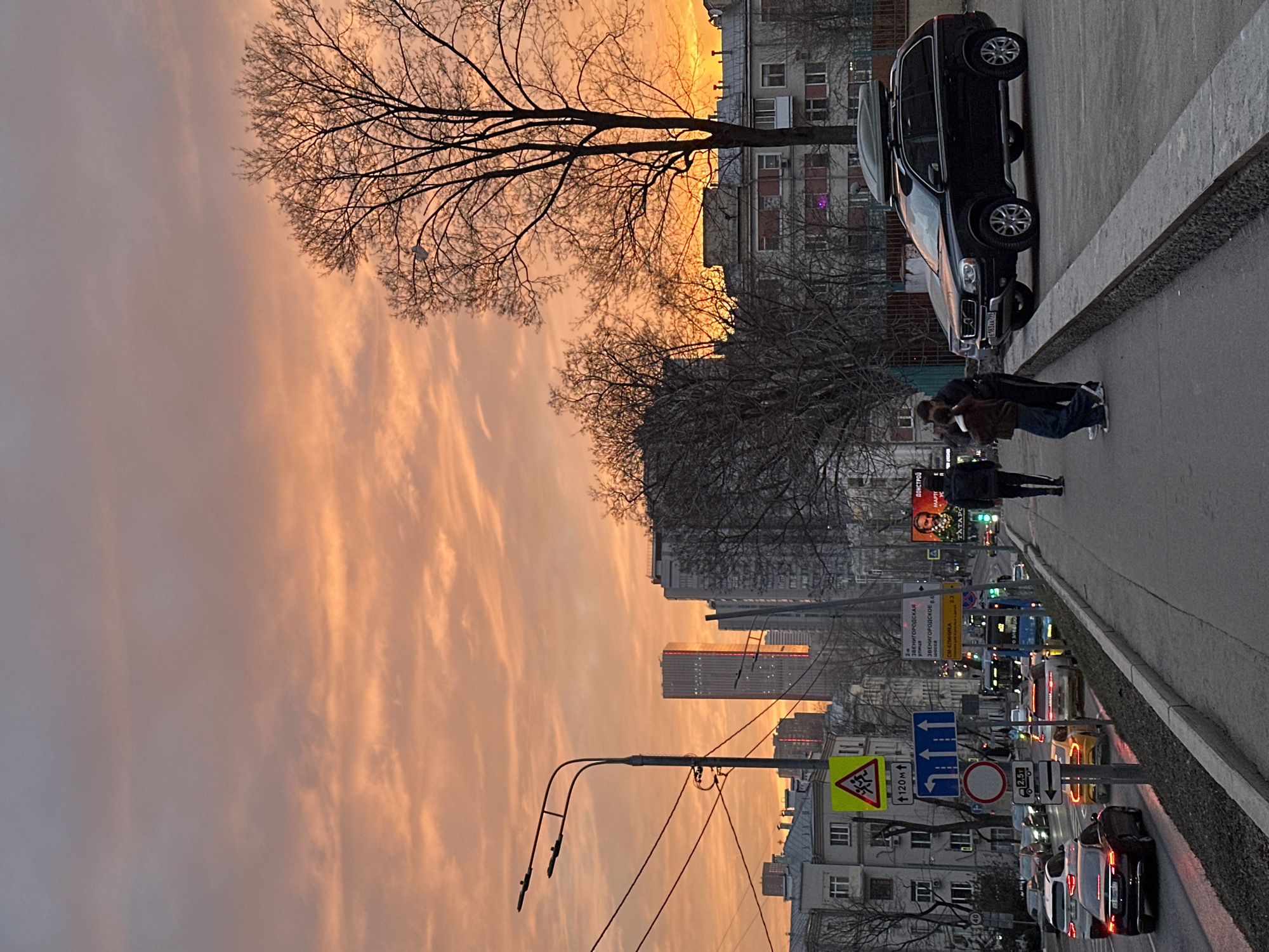 The image depicts an urban street scene in Moscow, Russia, at what appears to be either sunrise or sunset, characterized by an expansive sky displaying vibrant orange, pink, and purple hues with scattered cloud formations.
The scene is centered on a multi-lane road, identified by a visible street sign displaying "2-я Звенигородская улица" (2nd Zvenigorodskaya Street) and "ЗВЕНИГОРОДСКОЕ ШОССЕ" (Zvenigorodskoye Shosse), indicating the specific location within Moscow.
In the foreground, a concrete sidewalk extends from the lower left towards the mid-ground. To the left of the sidewalk, a black street lamp post with multiple fixtures and overhead power lines is present. Beneath it, a cluster of traffic signs includes a yellow triangular "Children" crossing sign with "120м" (120 meters) below, a blue rectangular sign indicating lane configurations (left turn and two straight lanes), a red circular "No Entry" sign, and a white rectangular sign displaying a truck icon with "2.5т" (2.5 tons) and an arrow.
The road itself is occupied by moderate vehicular traffic, with several cars, including sedans and SUVs, whose taillights are illuminated. A blue bus is visible in the middle distance. Traffic lights further down the road show green signals.
On the right side of the image, two individuals are positioned on a paved area adjacent to the sidewalk, near a parked black Volvo XC90 SUV, which is equipped with a roof cargo box and displays license plate "E183AA 799". One person, wearing a brown jacket and blue jeans, faces away from the camera, leaning towards a second person, dressed in a dark jacket and trousers, who is partially obscured. A third individual, wearing a dark jacket with light-colored striped pants, walks away from the camera on the sidewalk.
The environment includes several bare-branched trees, indicative of a dormant season. In the background, a mix of architectural styles is present: older, multi-story residential buildings are visible on the left, while prominent modern skyscrapers, including one with distinctive vertical red illuminated lines (likely the "Imperial House" building), dominate the central and right background skyline. A large digital billboard on the right side of the road displays an image of a historical figure, possibly Karl Marx, along with Cyrillic text, potentially "МАРТ" (March).
