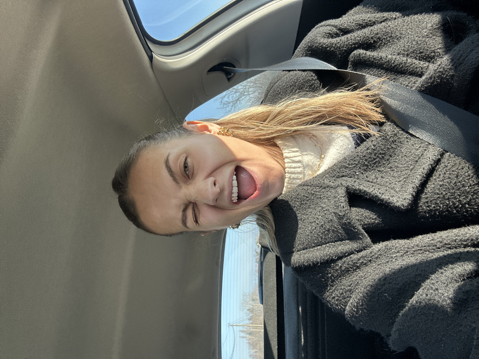 A young adult female is depicted seated inside a vehicle, with the scene identified as occurring in Saint-Cyprien, France. The subject, with blonde hair and a light complexion, is centrally positioned and directly facing the camera. Her left eye is closed in a wink, and her mouth is wide open, revealing both upper and lower teeth and her tongue. She is wearing two gold-colored, intricate earrings. Her attire consists of a cream-colored ribbed knit sweater, overlaid with a dark, textured, wide-lapel coat. A thin gold necklace with a small pendant is visible on her neck. A black seatbelt is fastened across her left shoulder and chest. The vehicle's interior headliner is light gray. Through the rear window, a clear blue sky is visible above a distant landscape featuring bare trees or dormant vegetation and a utility pole with power lines on the left. The scene is brightly illuminated, indicating a sunny day.