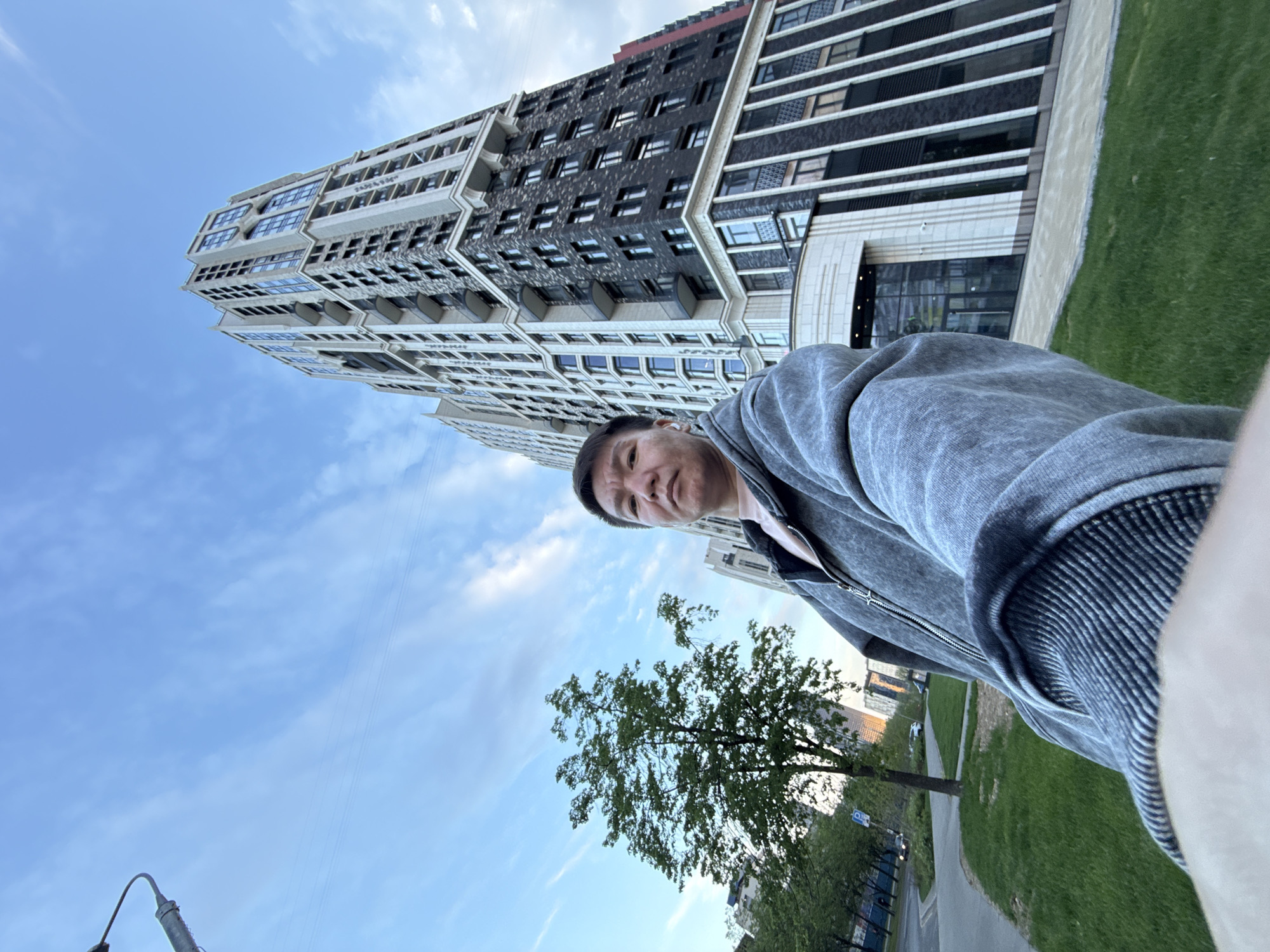 A man is taking a selfie outdoors. He is wearing a gray zip-up hoodie. Behind him is a tall building with a modern design, with light and dark panels. A tree is visible in the lower part of the frame, and a sidewalk runs alongside a patch of grass. The sky is blue with scattered clouds. Based on the architectural style of the building, the location is likely within the city of Khimki, Russia.